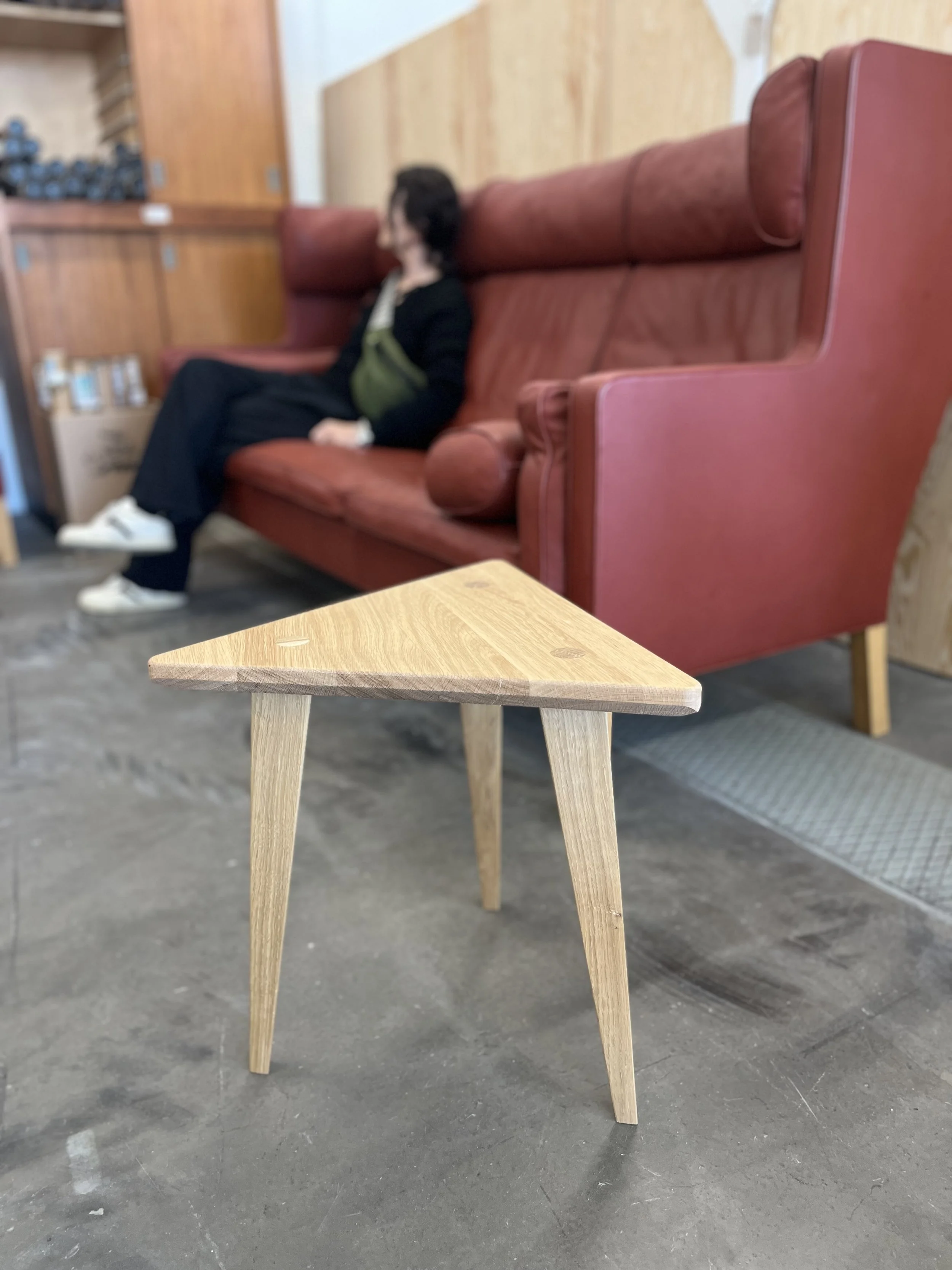 Ett litet trämöbel med tre ben placerad på en betonggolv, i bakgrunden finns ett rödbrunt soffbord och en vägg med träpanel.