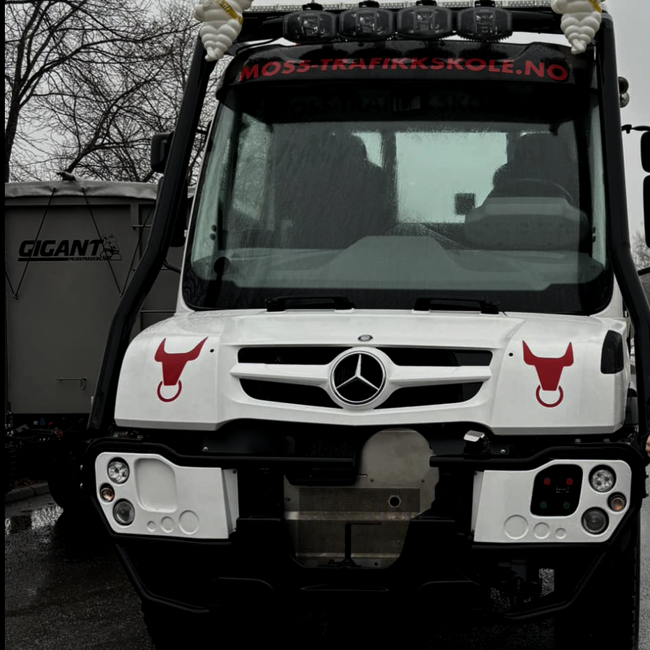Et frontbilde av en hvit lastebil med Mercedes-Benz-logo, dekorert med røde hundetemperamenthoder på begge sider, og en nettsideadresse med tekst 'MOSS-TRAFIKKSKOLE.NO' øverst foran på førerhuset.