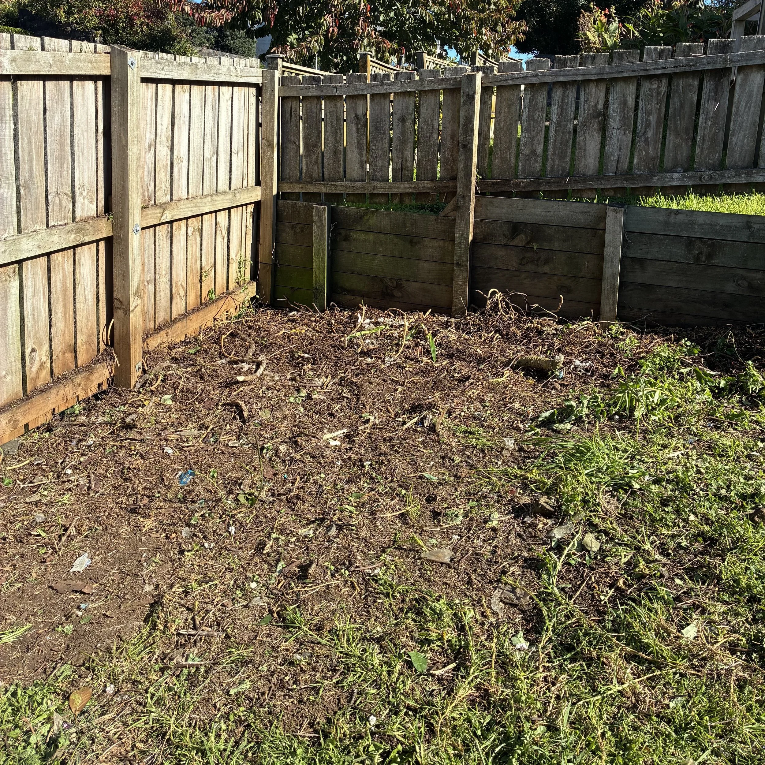 A wooden fence with vertical planks, partially broken, in an outdoor yard with soil, small plants, and grass. There are some trees and bushes beyond the fence.