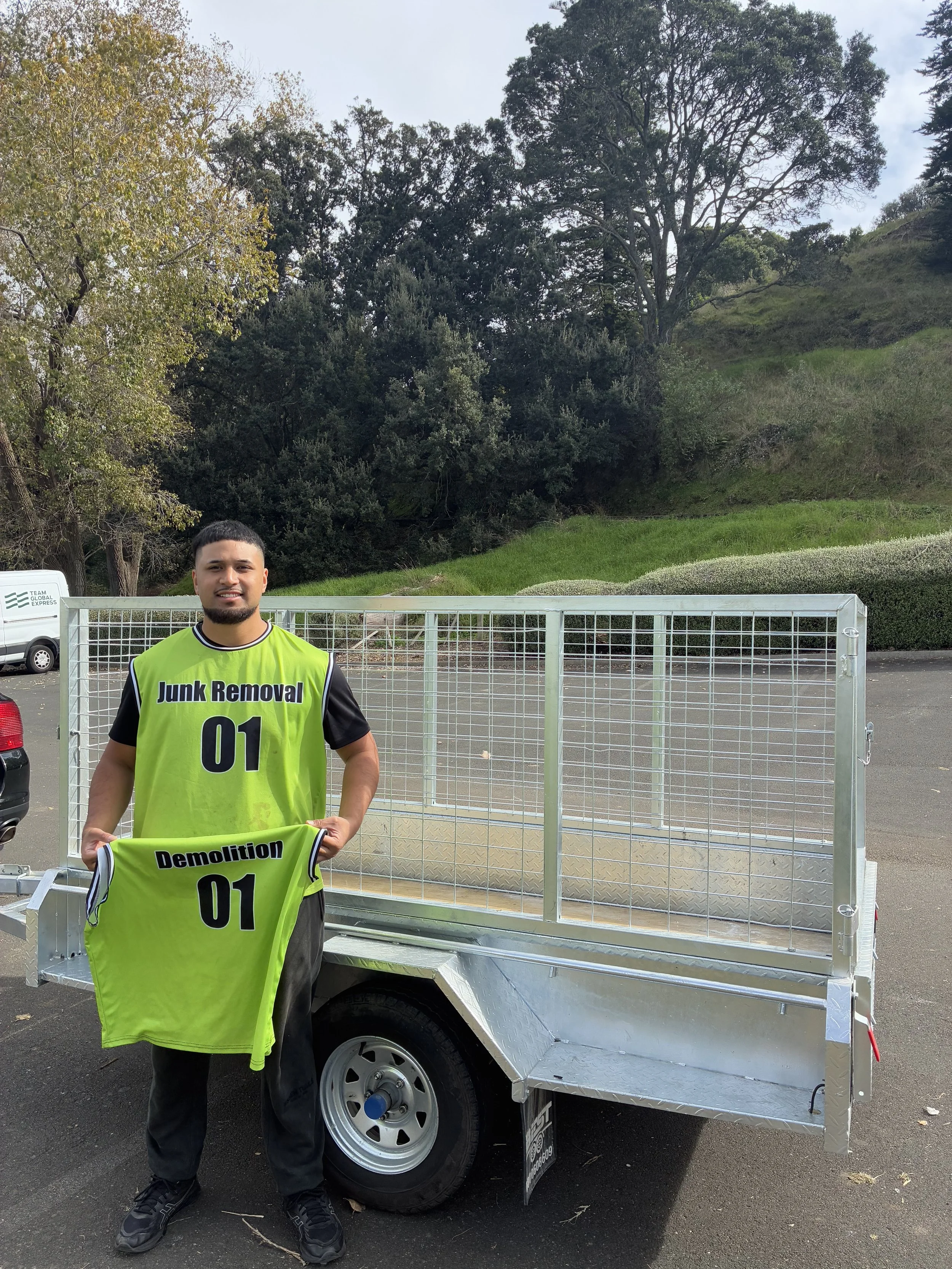 A man wearing a bright yellow-green vest with the words "Junk Removal" and the number "01" is standing in front of a small utility trailer. The trailer is empty and parked on asphalt, with a backdrop of trees and grass on a cloudy day.