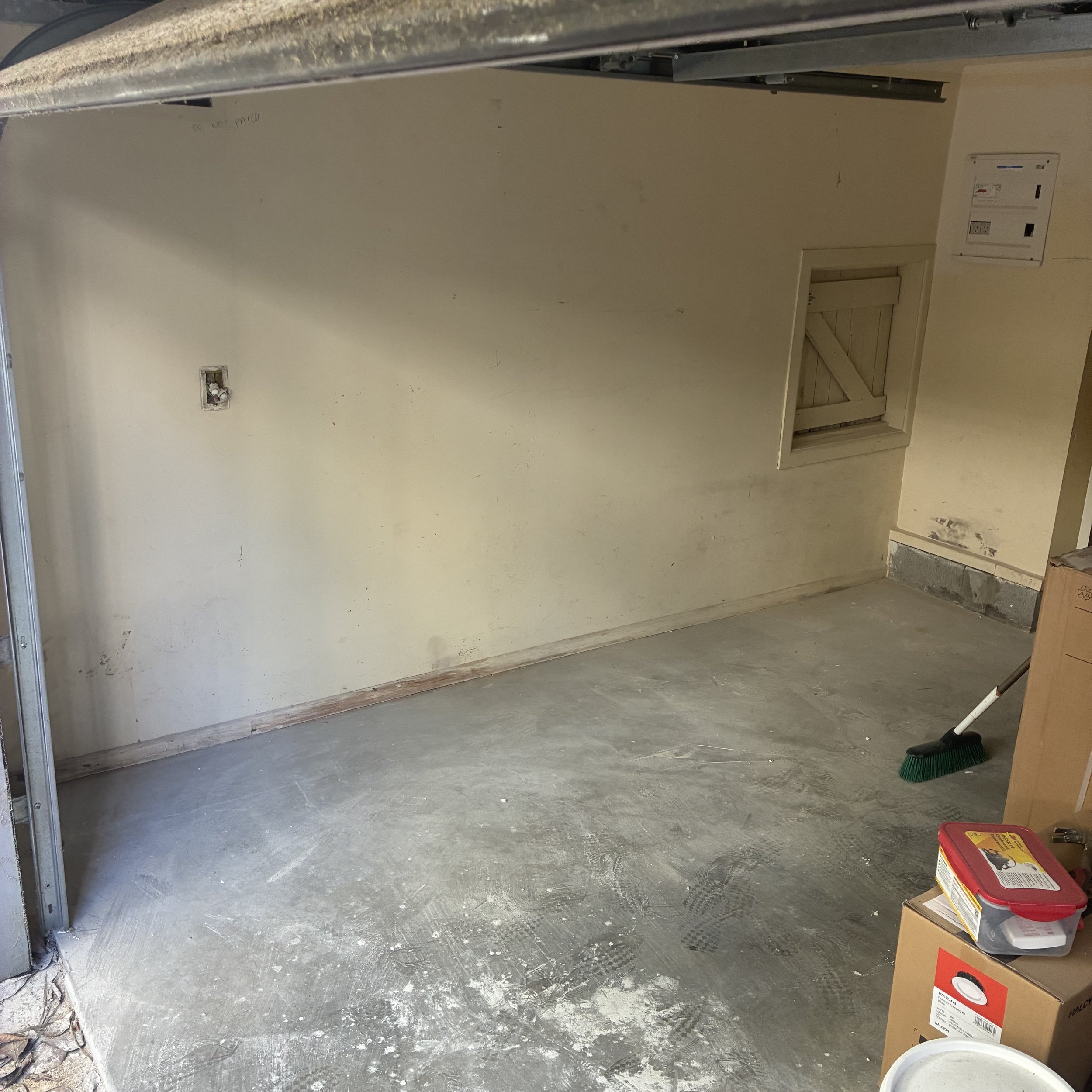 Empty garage with unfinished concrete floor, some boxes, a broom, and an electrical panel on yellow wall.