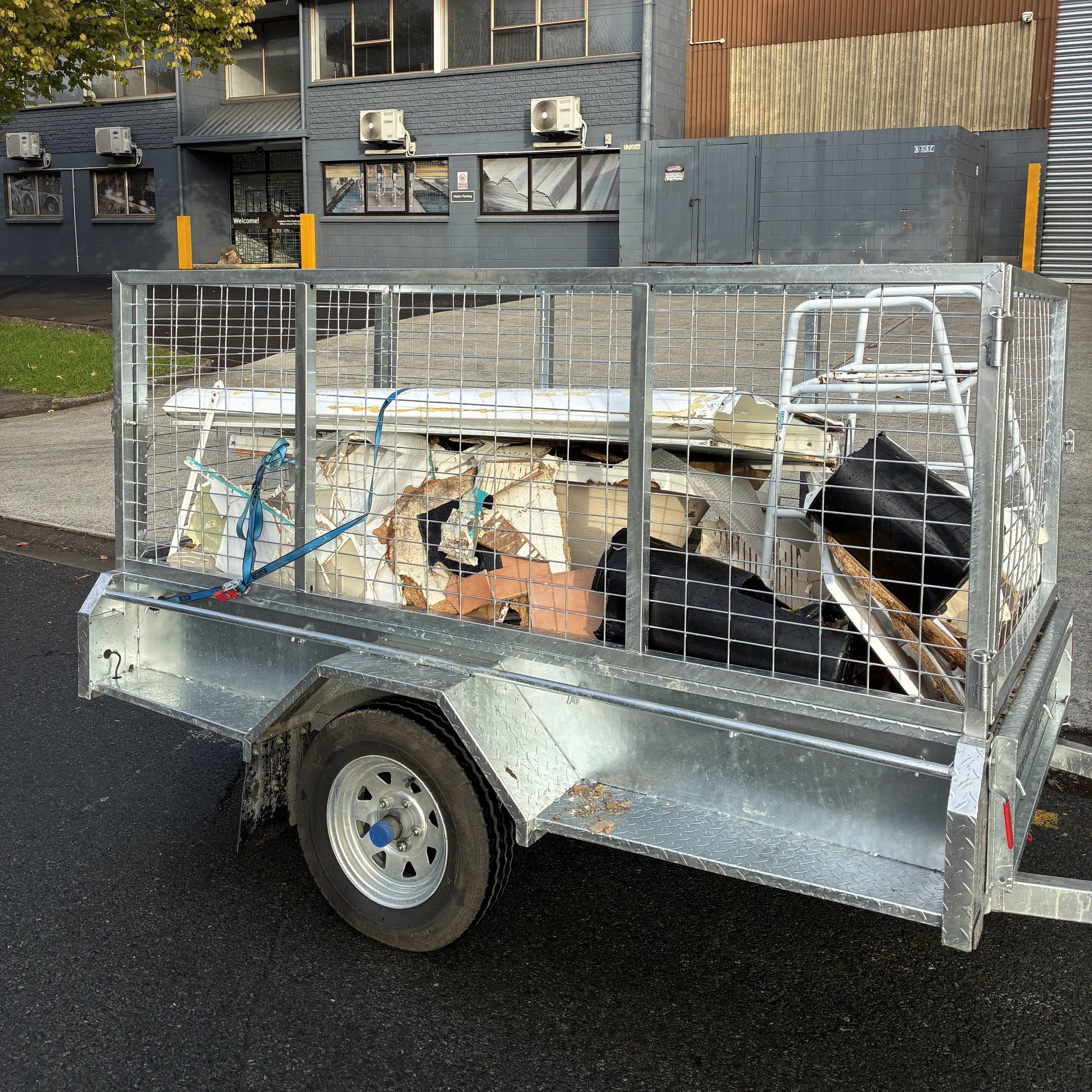A small cargo trailer attached to a vehicle, carrying discarded debris including broken wood, roofing material, and other trash, parked on a street in front of a commercial building.