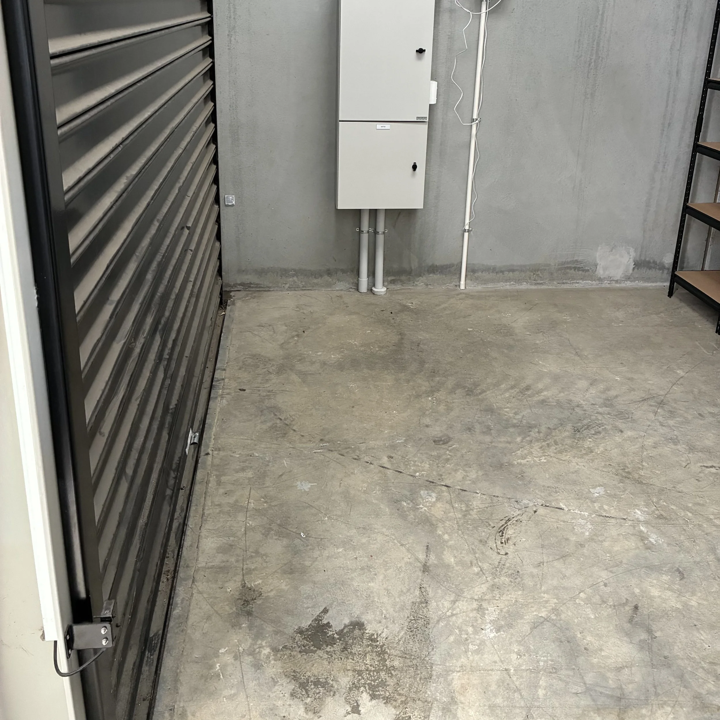 Empty concrete floor with a gray wall, a black metal shelf on the right, a gray electrical panel on the wall, and a section of a gray metallic tool cabinet on the left.