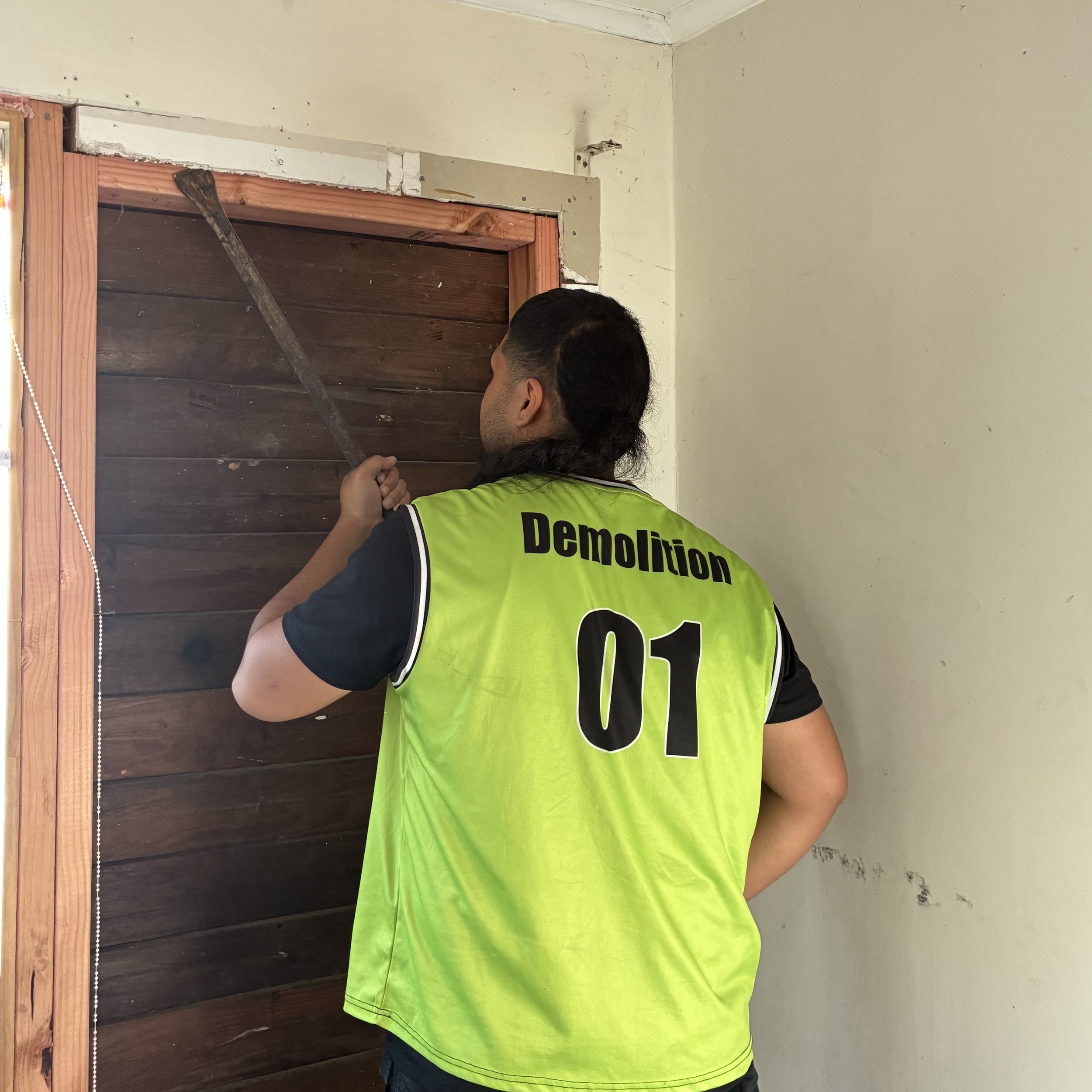 A person with dark hair wearing a yellow safety vest labeled "Demolition" working on a door frame, holding a metal crowbar, with a wood-paneled door and a white wall in the background.
