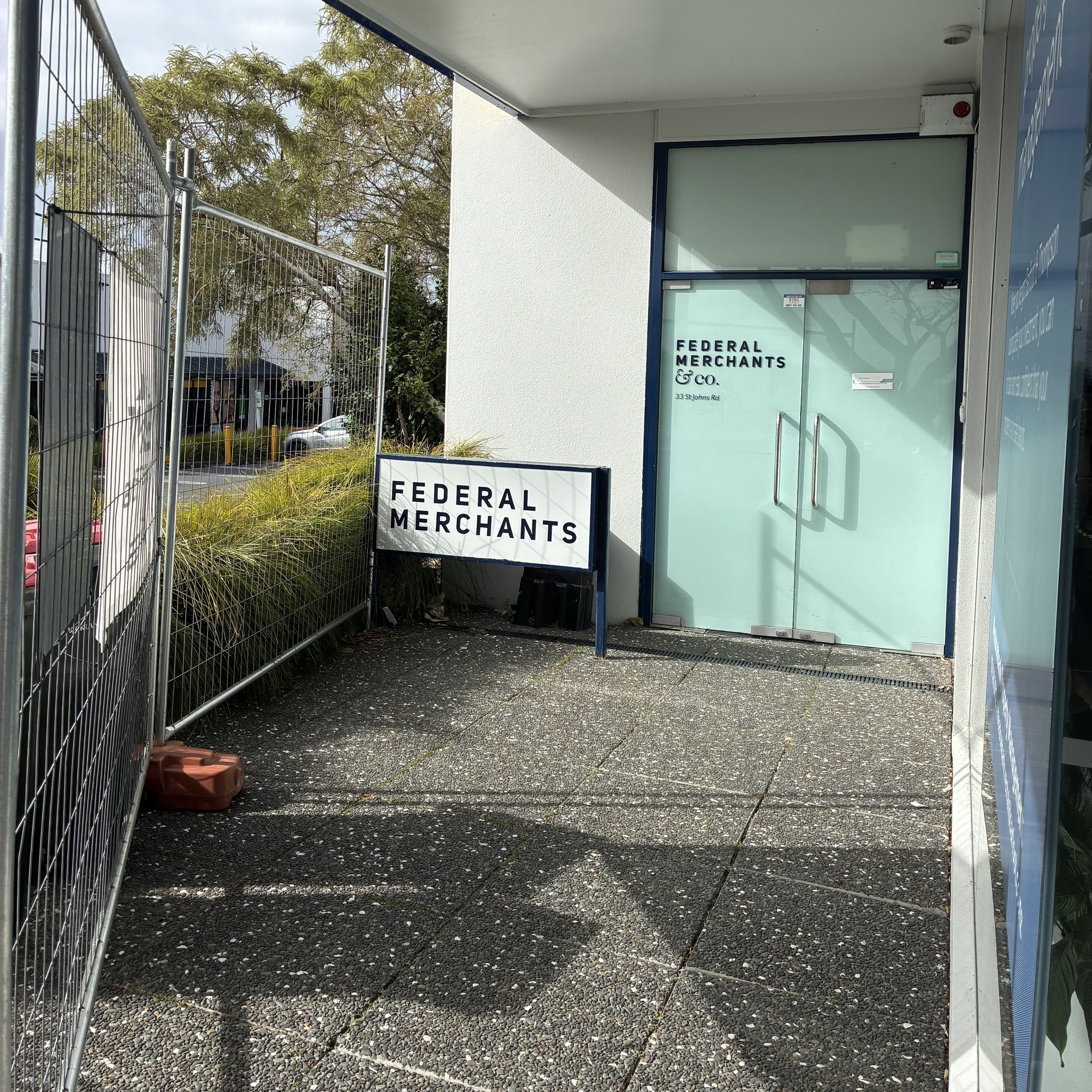 The exterior entrance of a small commercial building labeled 'Federal Merchants & Co.' with glass door and sign, metal fencing on the left, trees and parking lot in the background, and concrete sidewalk in the foreground.