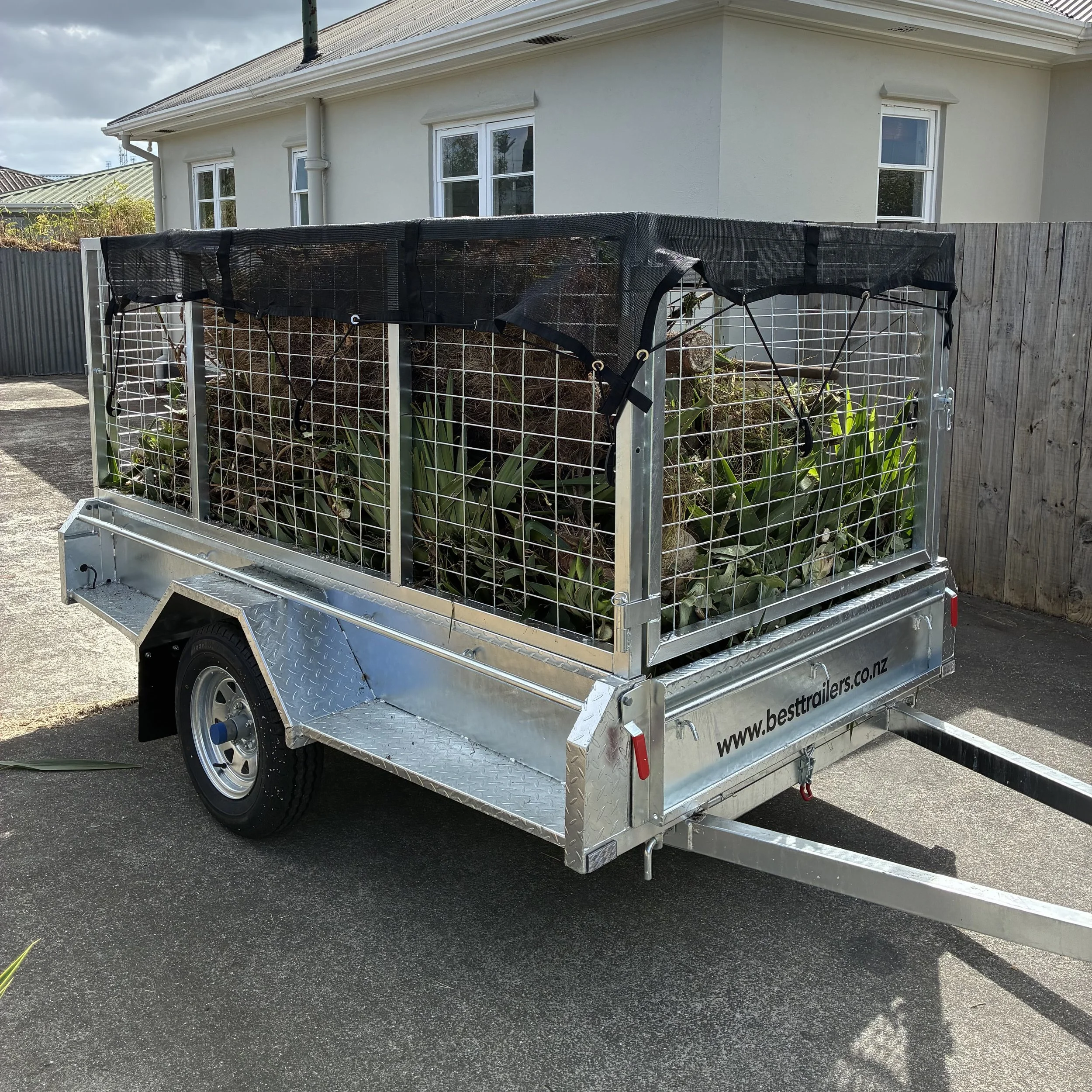 A small enclosed trailer with a metal frame and mesh sides parked on a paved driveway in front of a house. The trailer contains plants or soil and is situated near a wooden fence.
