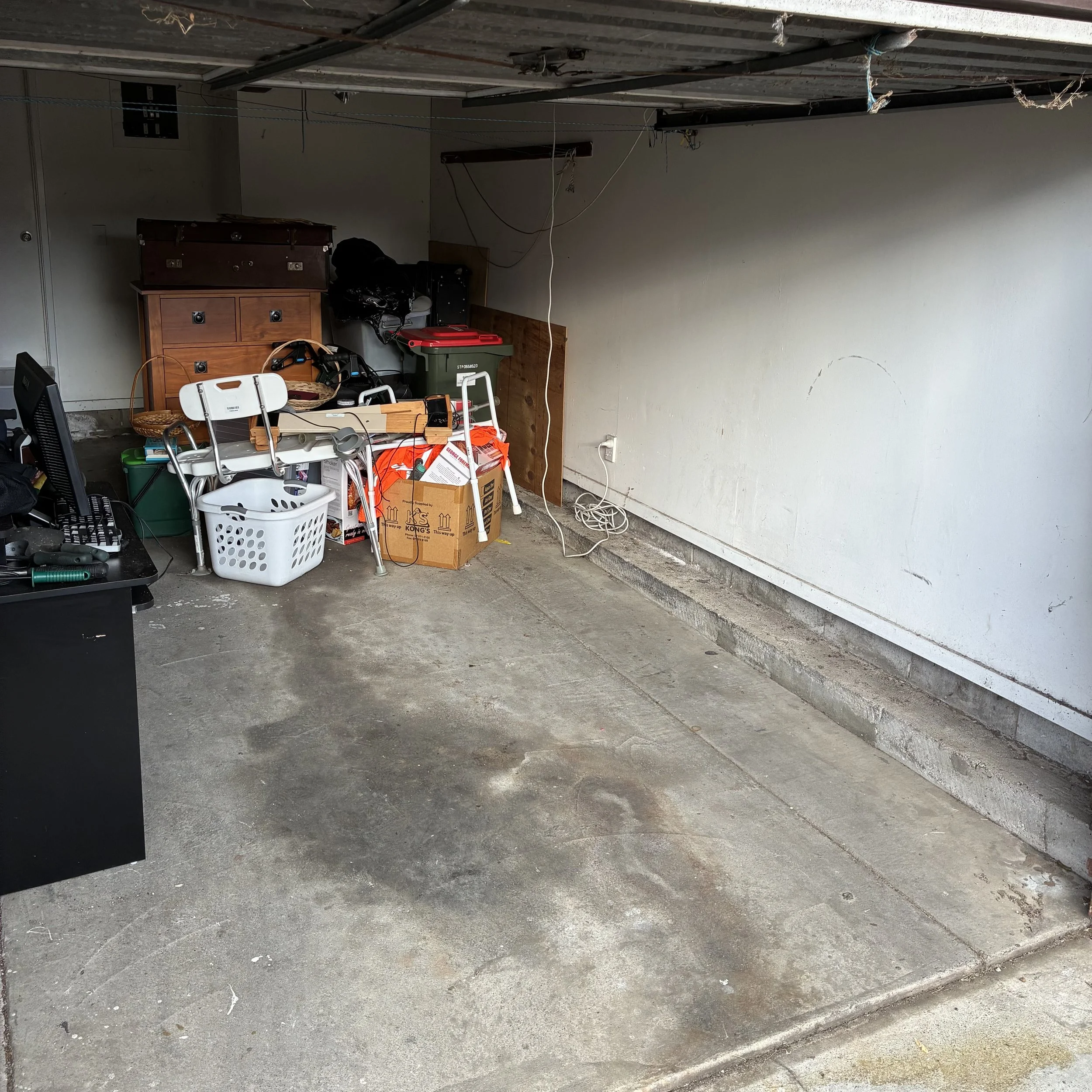 A cluttered garage with various items including baskets, boxes, furniture, and miscellaneous objects scattered on the floor and against the wall.
