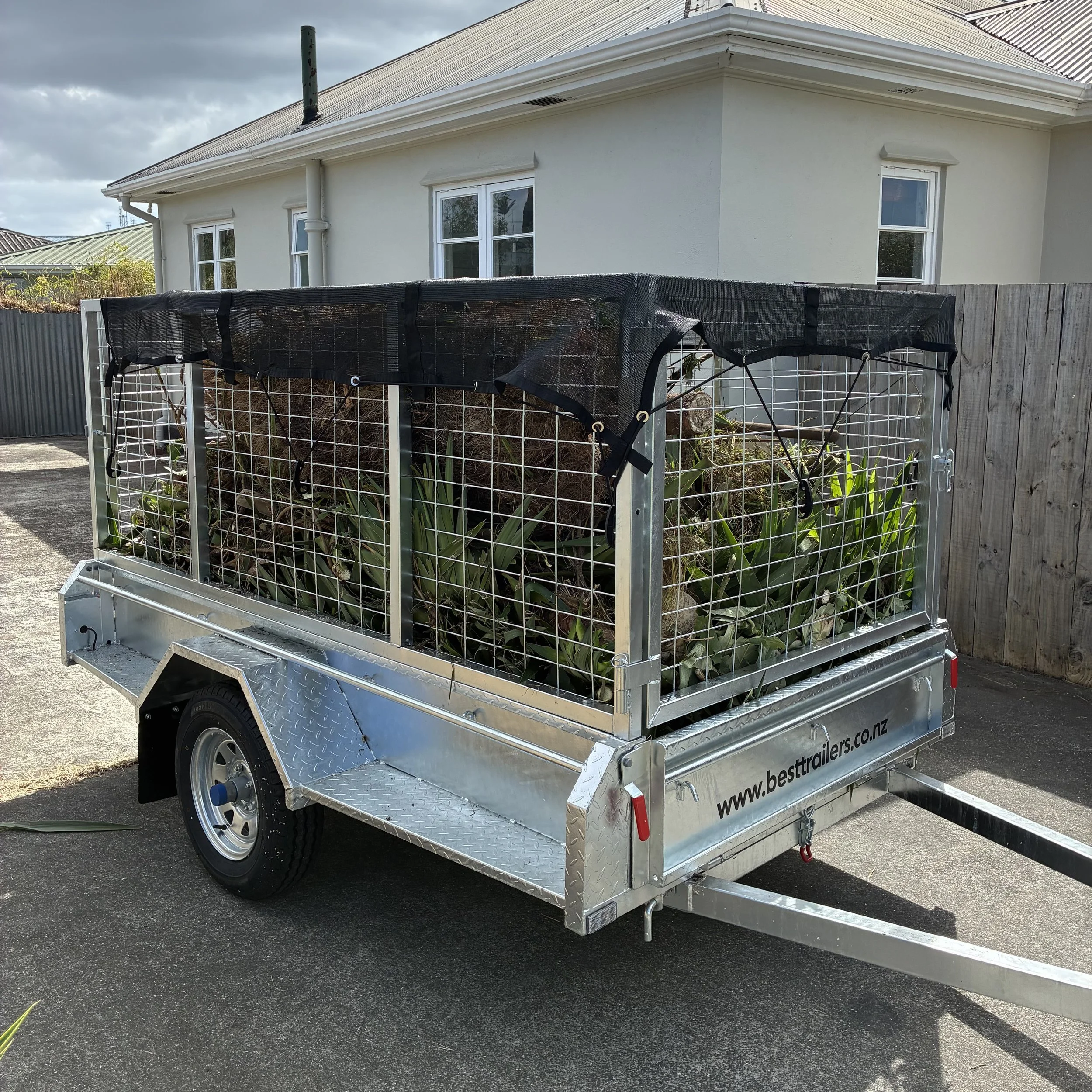 A metal trailer on a paved driveway, filled with soil and plants, with a house in the background under a cloudy sky.