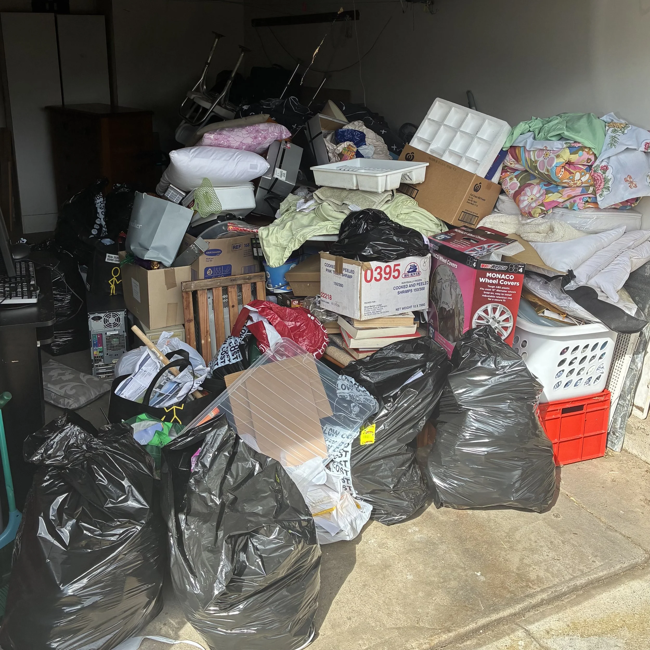 A cluttered garage with various items and trash bags piled up, including boxes, bedding, and household items.