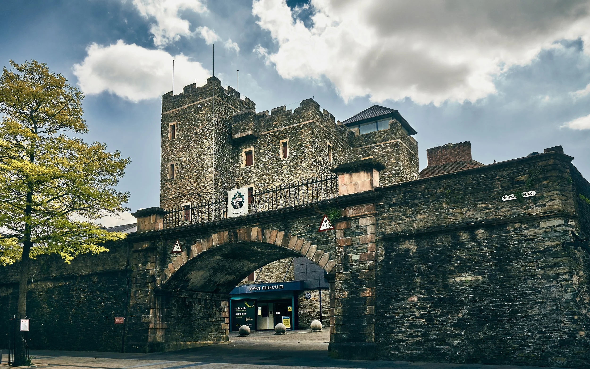 View of a historic stone castle-like tower with battlements, small windows, and a modern glass and wood section on top, situated above an arched stone entrance with a sign that reads 'tower museum.' There is a tree with green leaves on the left side and a cloudy sky in the background.