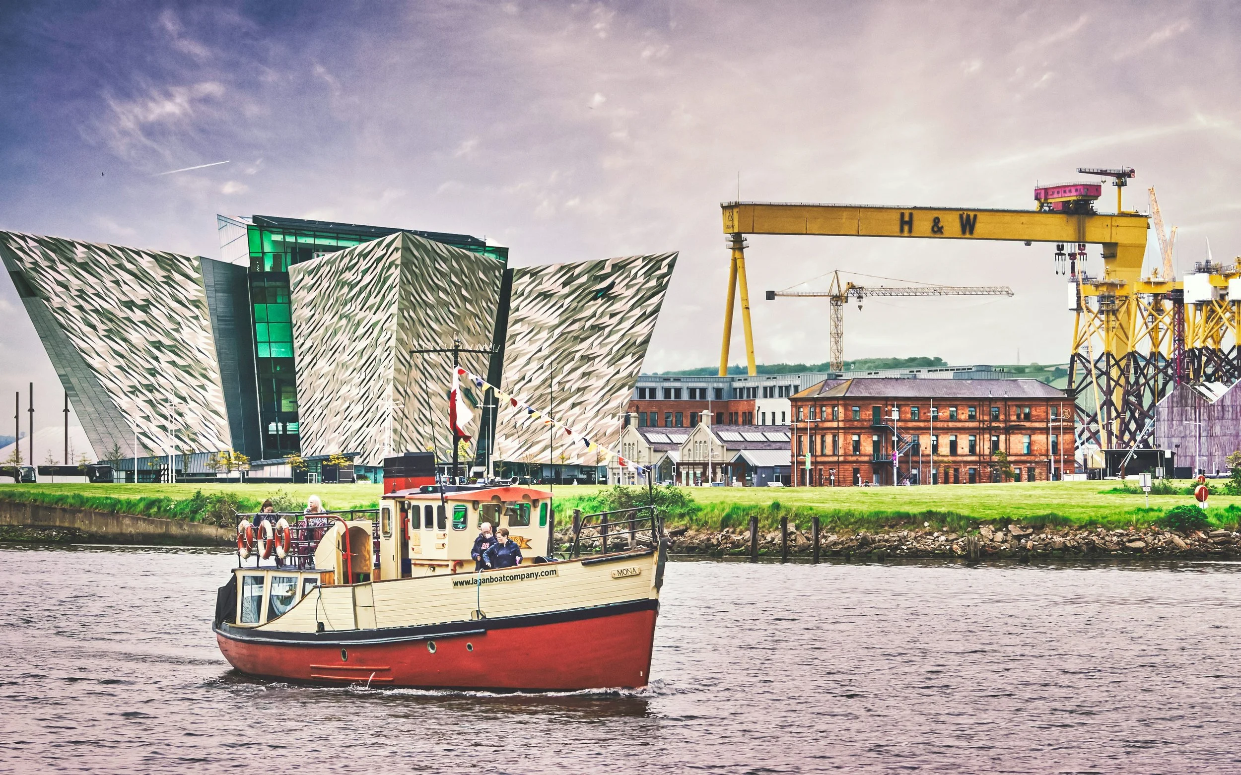 A boat on a river with modern city buildings and construction cranes in the background, under a cloudy sky.