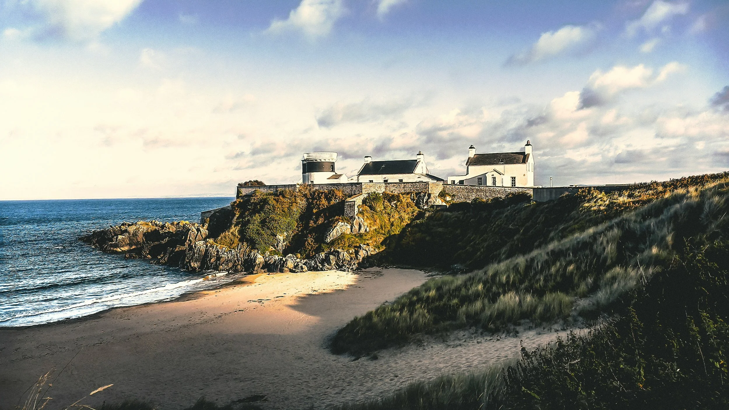 A coastal scene with a sandy beach and rocky cliffside, featuring a white lighthouse and buildings atop the cliffs. The ocean is visible to the left, and the sky is partly cloudy.