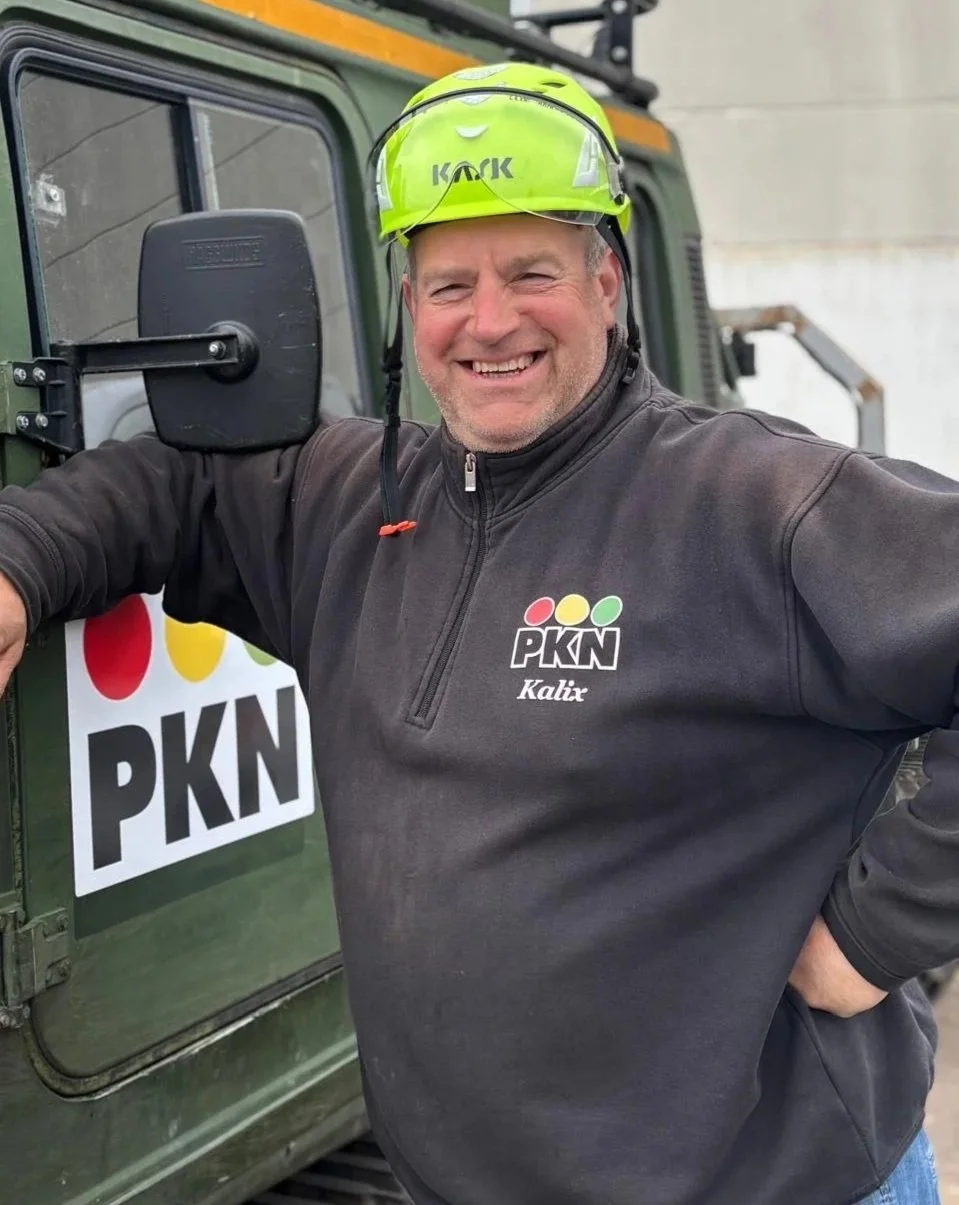 Glad man i svart jacka och grönt skyddshjälm står bredvid en grön fordon, som har en logga med texten "PKN".