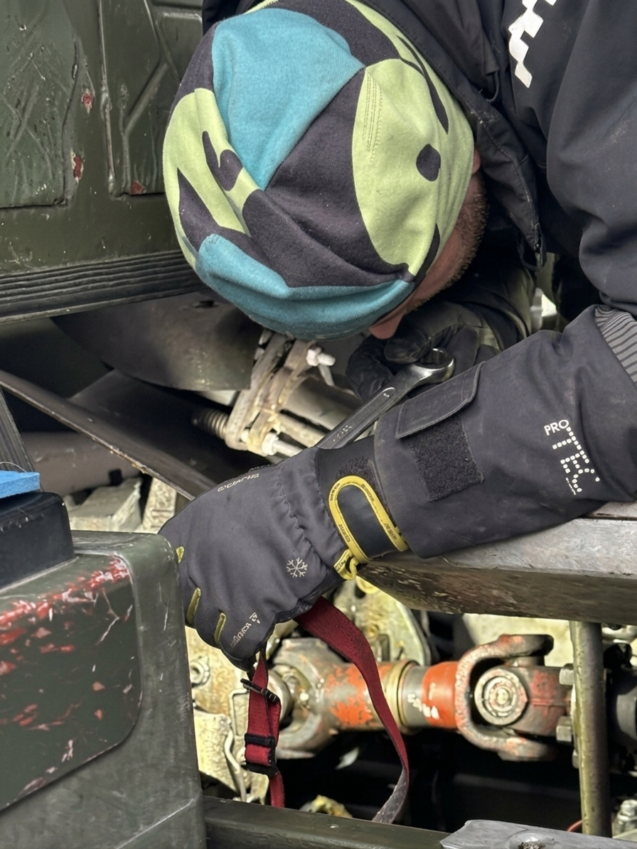 En person som arbetar under en motor eller maskin, använder svart handskar och har på sig en mössa med färgglad design.