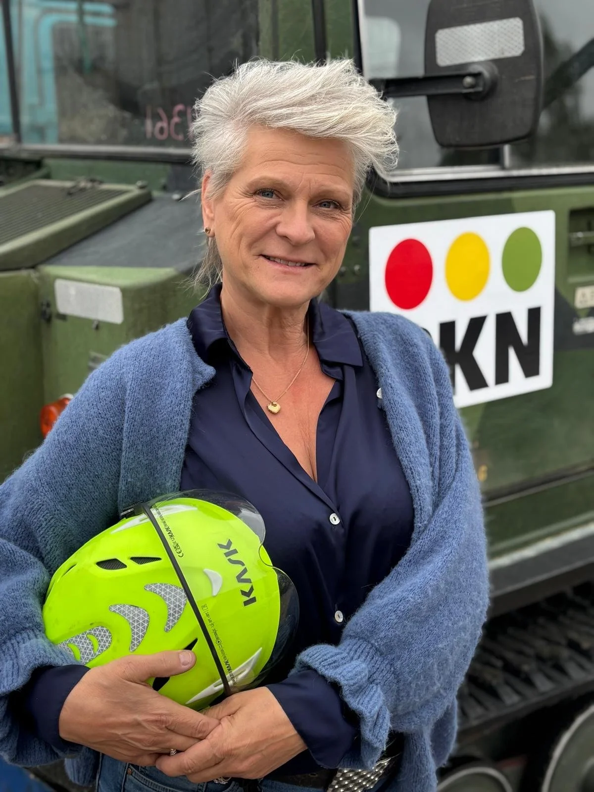 en äldre kvinna med grått hår som håller en ljusgrön cykelhjälm, står framför en militärbil med en logga som har tre färgade cirklar och bokstäverna KKN.