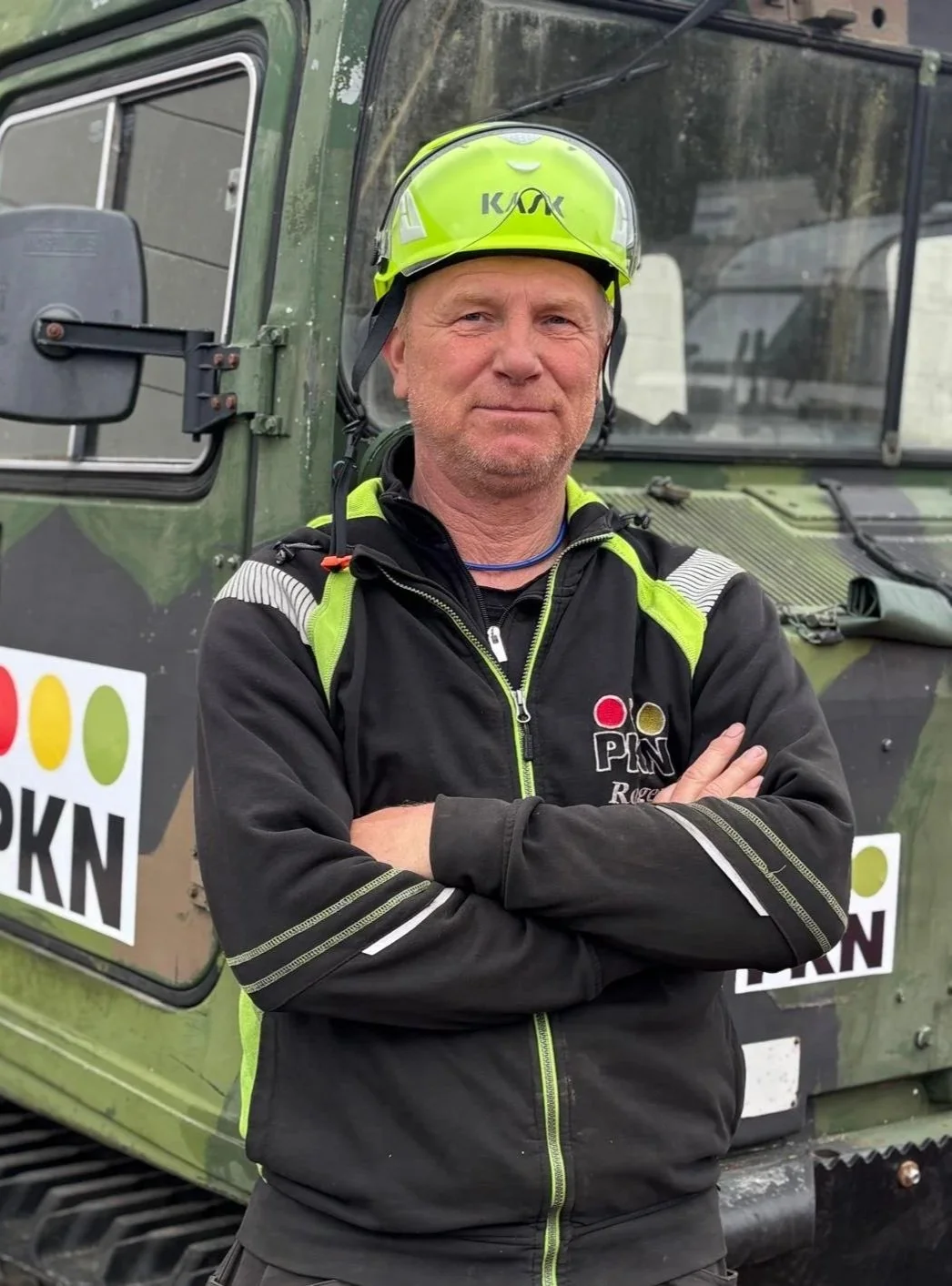 Man i rallykläder och hjälm står framför en grön terrängbil, arms cross, med logotypen PINK rally. Ser självsäker och nöjd ut.