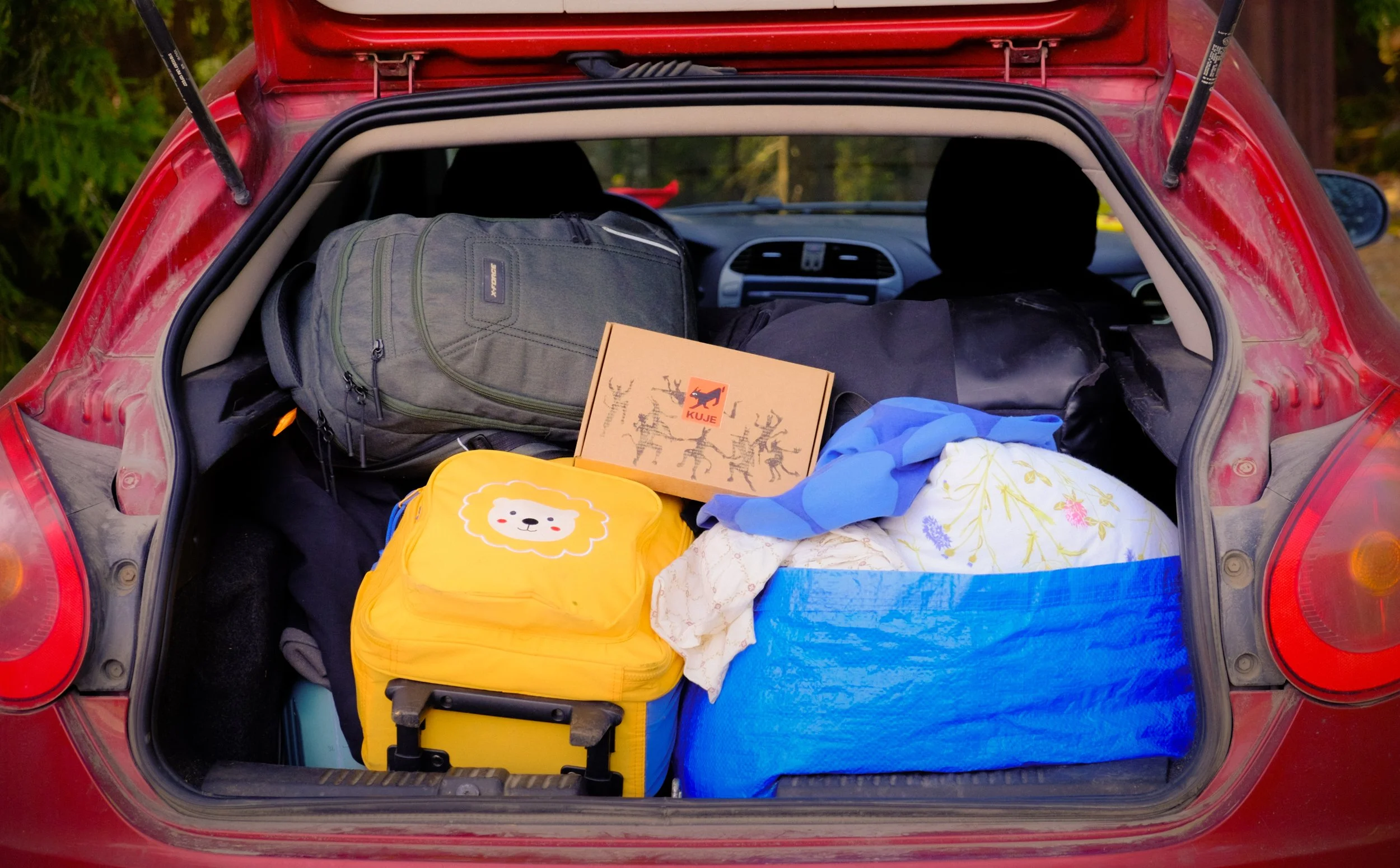 Punainen auto, jonka takakontissa on seikkailu laatikossa, matkalaukkuja, paketteja ja lastenvaatteita.