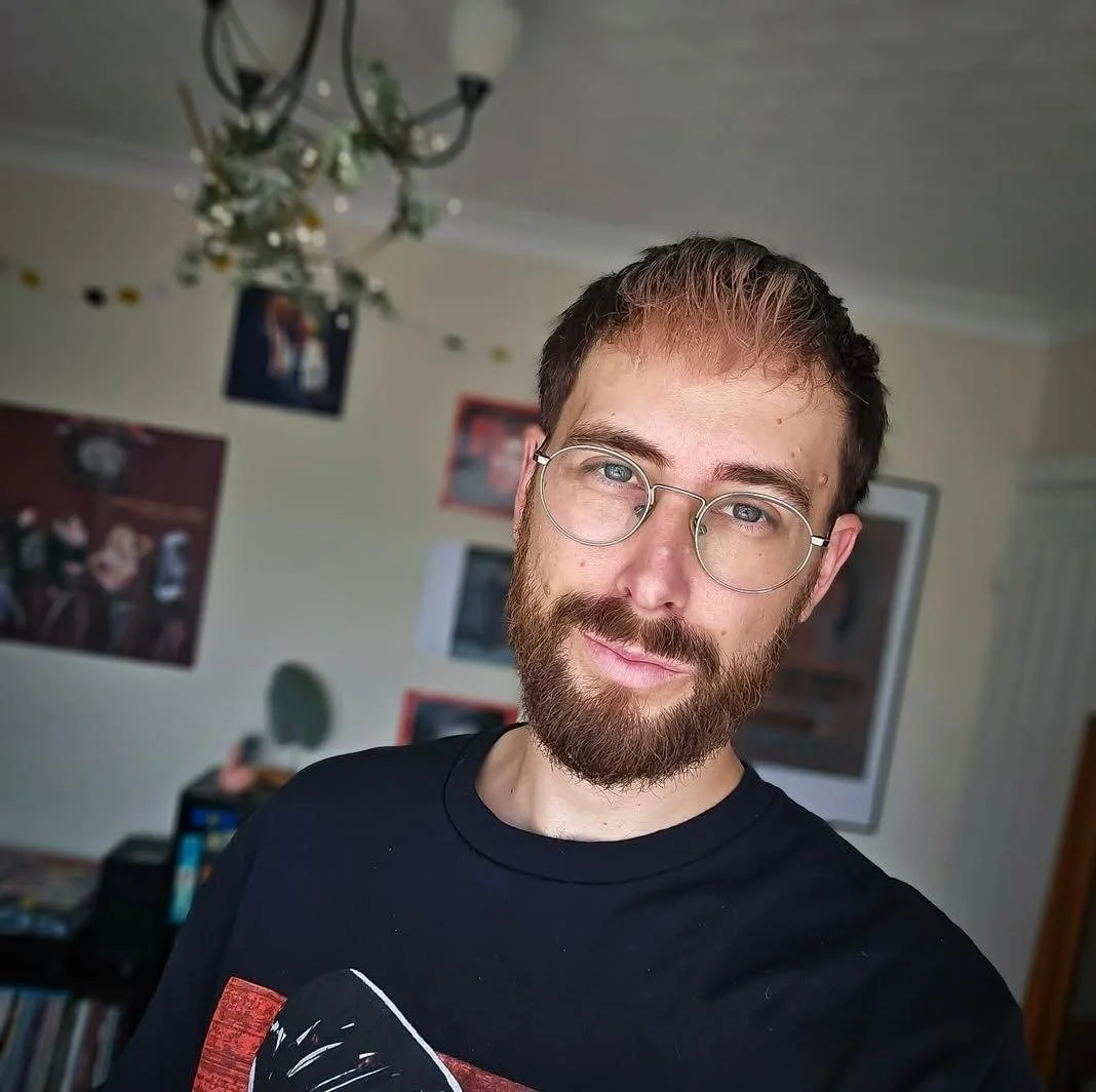 A man with glasses, a beard, and short brown hair taking a selfie in a room with posters on the wall and a chandelier hanging from the ceiling.