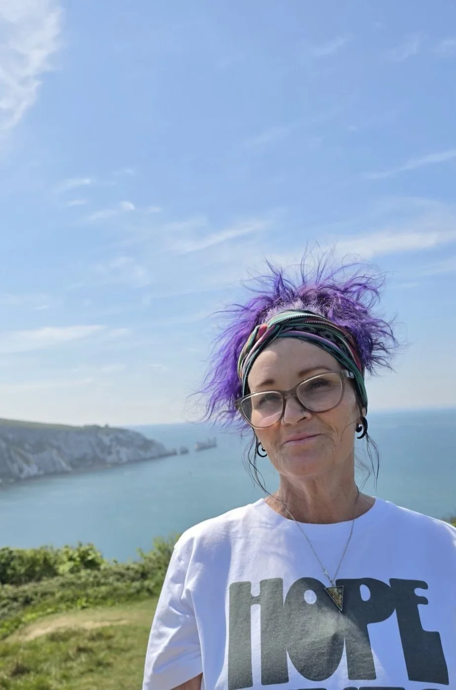 An elderly woman with purple hair, wearing glasses, earrings, a colorful headband, a necklace with a triangular pendant, and a white t-shirt with the word 'HOPE' on it, standing outdoors with a scenic view of water, cliffs, and a blue sky in the background.