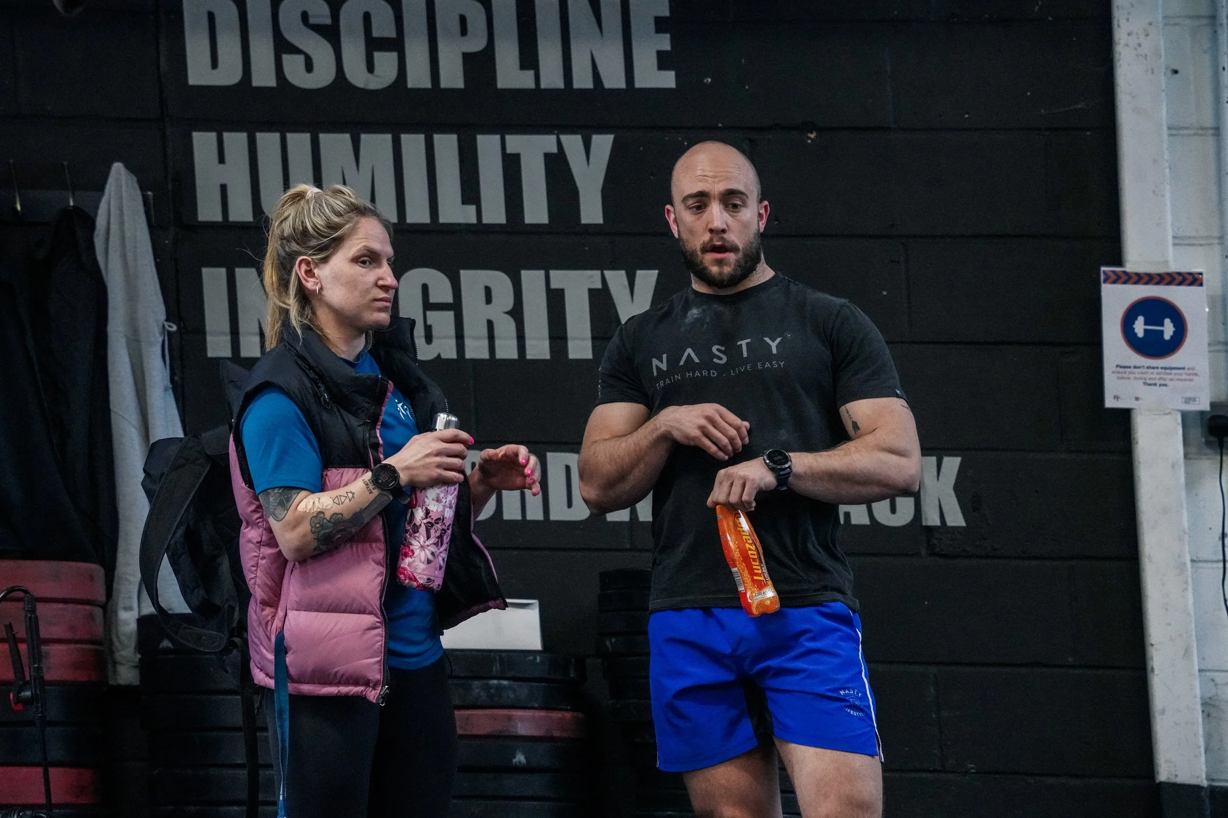 A man and woman in workout gear standing and talking in a gym. The background has a wall with large motivational words 'Discipline', 'Humility', and 'Integrity'. The man is holding an energy drink and wearing a black t-shirt with the word 'Nasty' on it, while the woman is holding a small bottle and wearing a pink vest over a blue shirt.
