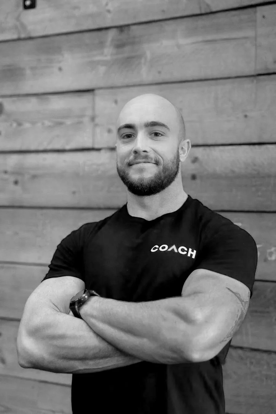 A muscular man with a beard and a shaved head, crossing his arms and smiling slightly, wearing a black shirt with the word 'COACH' printed on it and a watch, standing in front of a wooden wall.