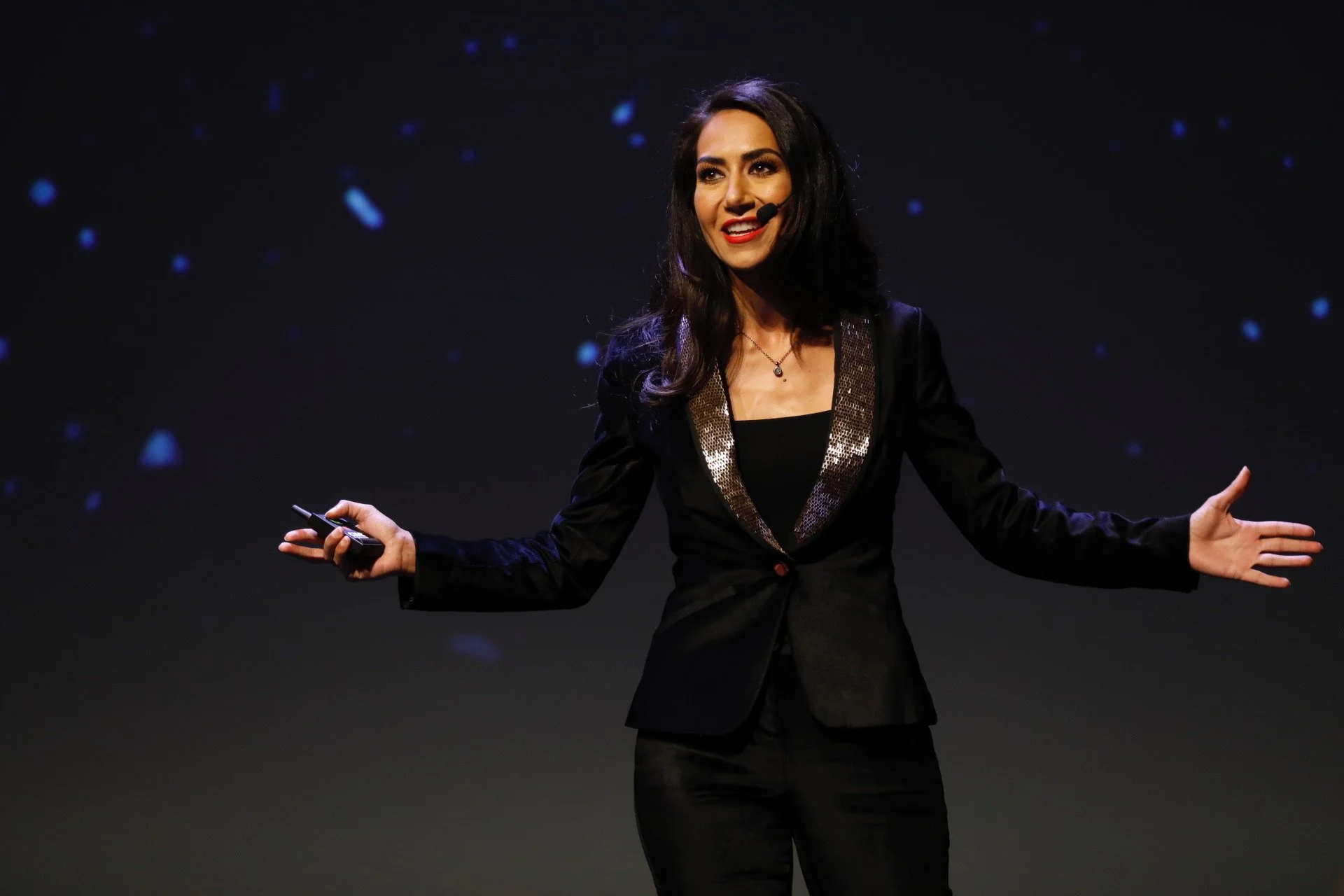 A woman wearing a black suit with gold sequin lapels giving a presentation on stage, gesturing with her hands, with a dark background and faint blue lights.