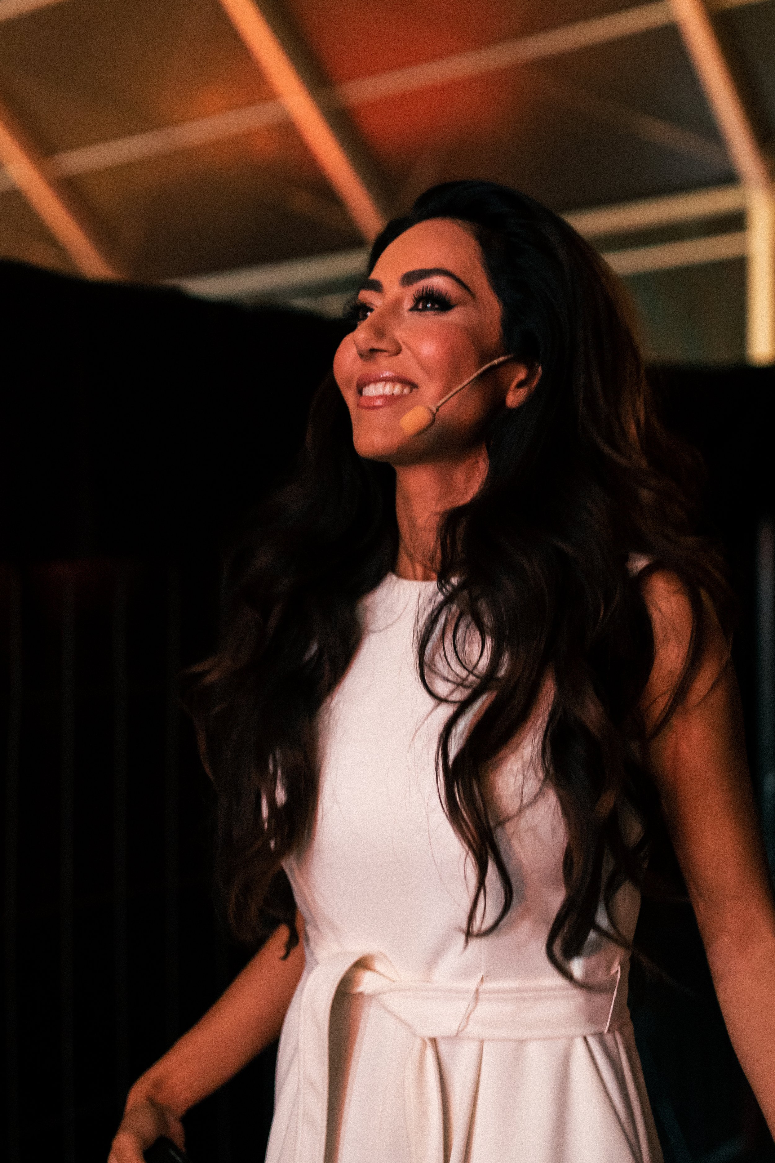 A woman with long wavy dark hair, wearing a white outfit and a headset microphone, smiling and looking to the side. The background shows a ceiling with exposed beams. in a reflective moment, neuroscientist and thought leader on the future of humanity