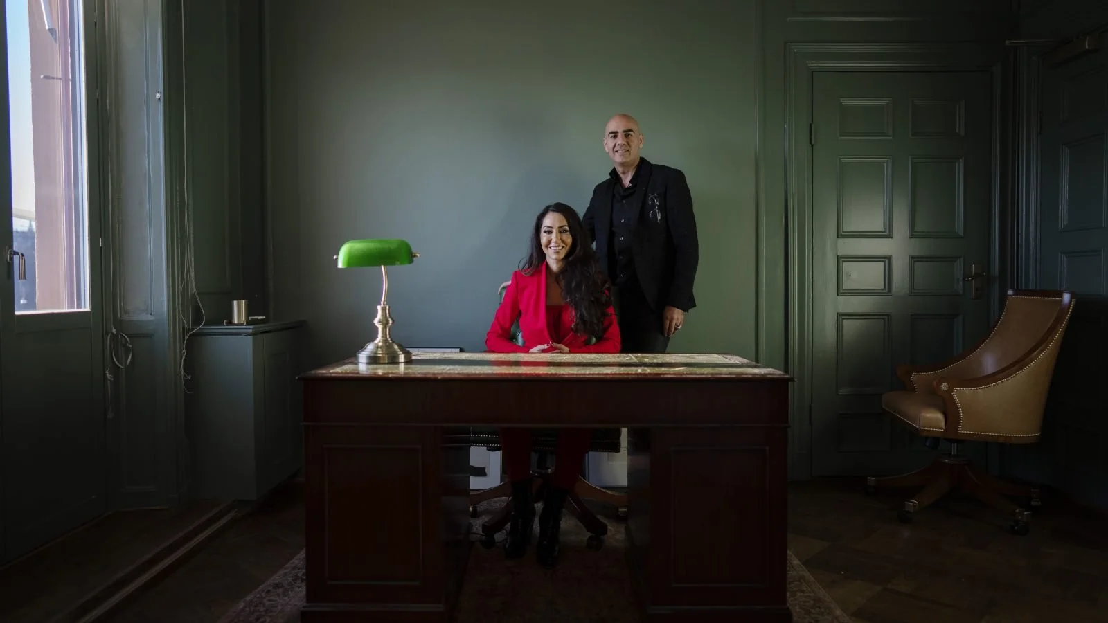 A woman sitting at a desk in a green-walled office with a green banker lamp, and a man standing behind her.