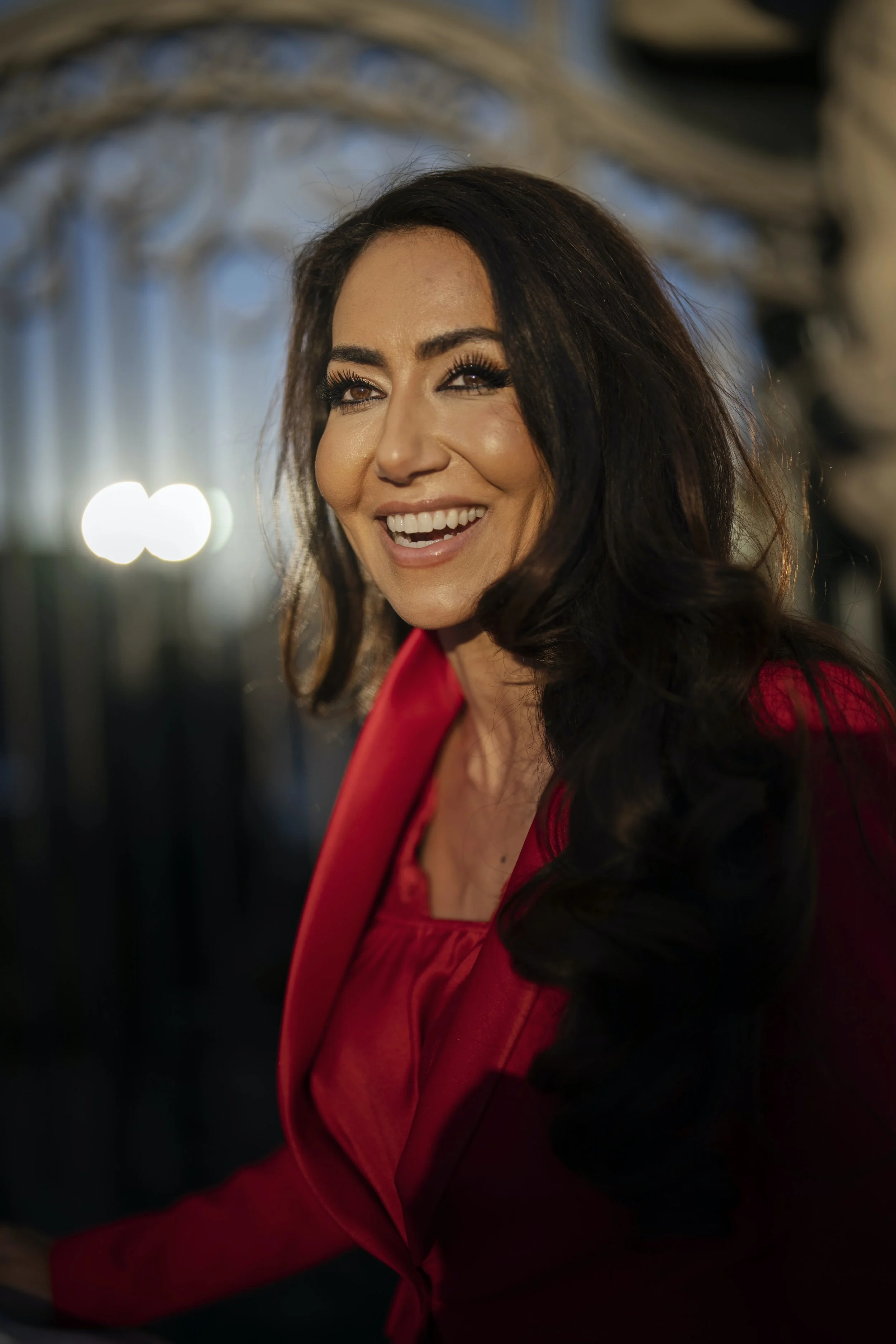 A woman with dark hair, wearing a red blazer, smiling and winking, standing in front of a blurred background with lights. Delivering a keynote on neuroscience and human potential at a major conference. Mouna Esmaeilzadeh speaking on stage about the n