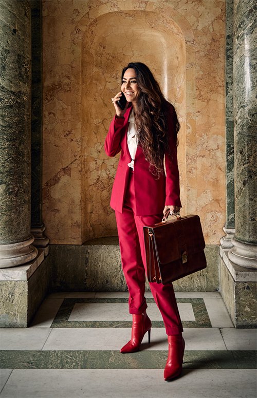 A woman in a red pant suit and matching red boots, speaking on her cellphone and holding a brown briefcase, standing in front of an ornate marble wall.