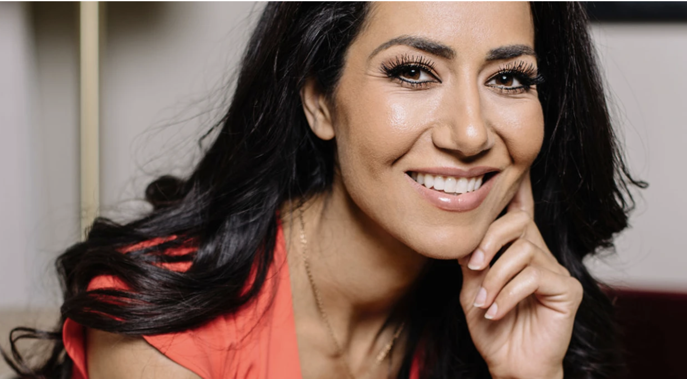 Close-up of a smiling woman with long dark curly hair, wearing a coral-colored top, resting her chin on her hand.
