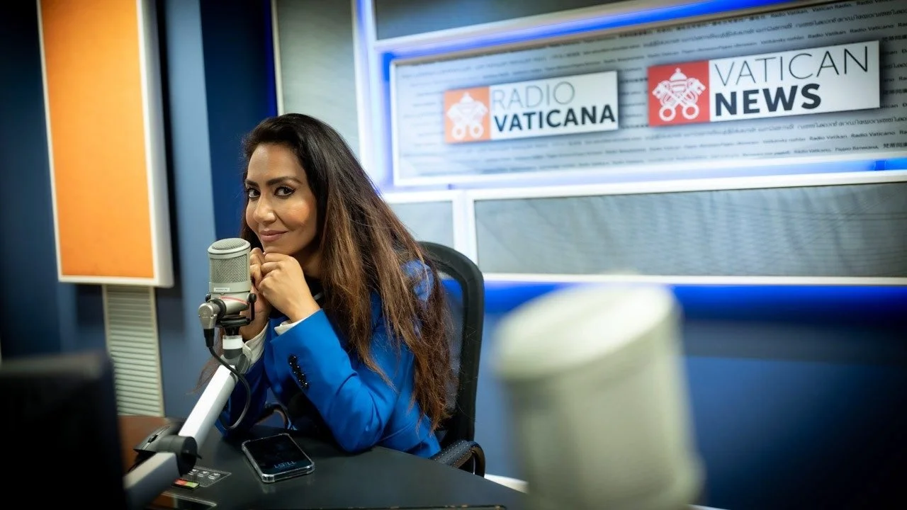 Woman in a blue blazer sitting at a radio station microphone in a broadcasting studio with Vatican-themed signs in the background.