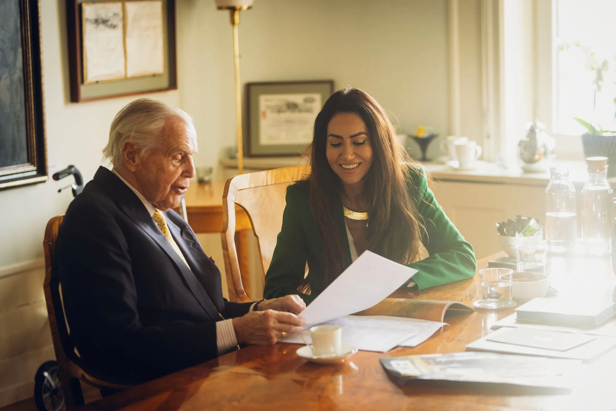 An older man and a woman sitting at a wooden table discussing papers. The man is in a wheelchair and dressed in a suit with a yellow tie, the woman is smiling, wearing a green blazer.