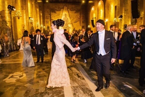 Couple dancing at a formal event in an ornate, gold-decorated ballroom with other guests dancing and socializing.