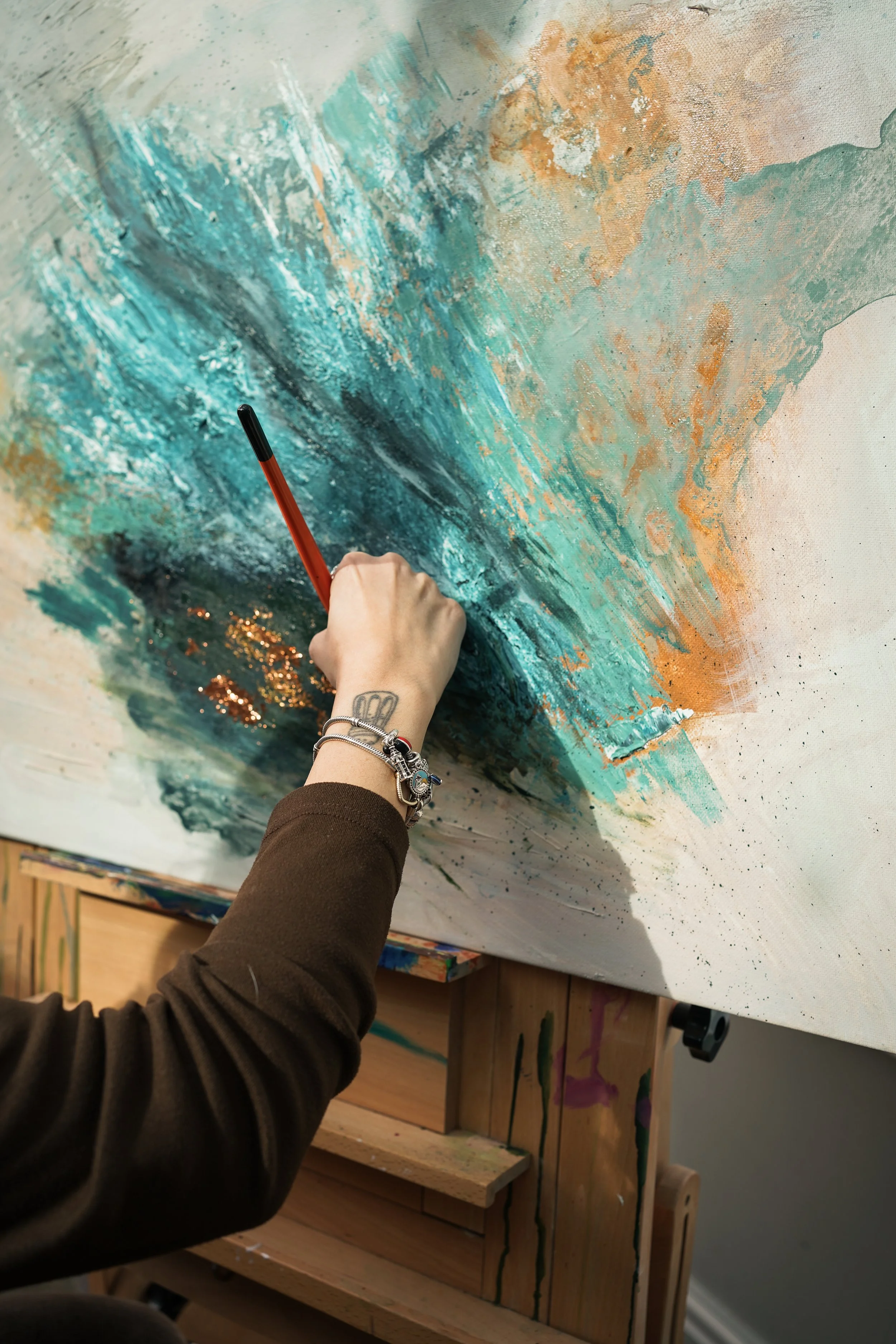 An artist's hand holding a paintbrush, creating an abstract painting with shades of blue, orange, and white on a canvas in an art studio.