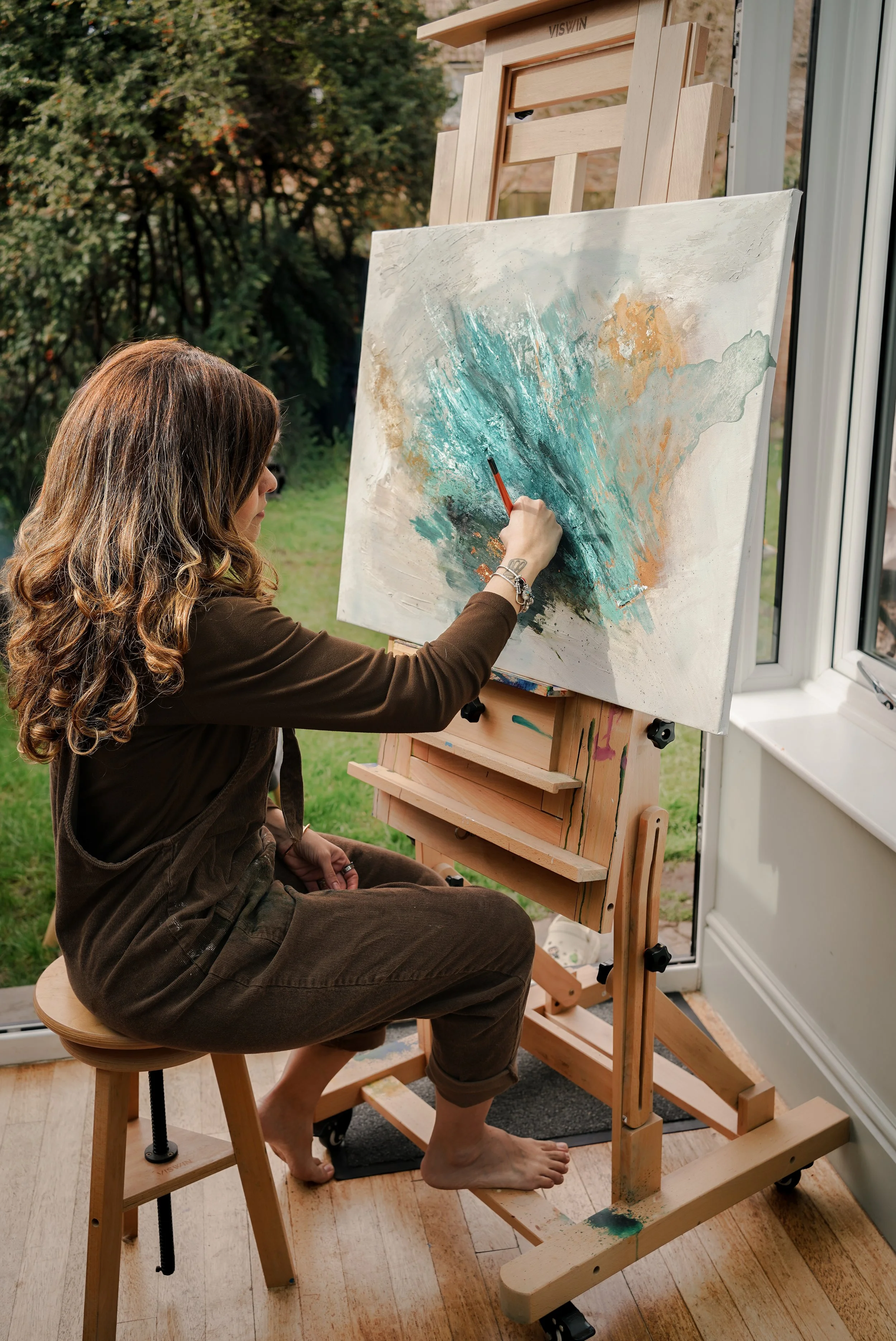 A woman with long, wavy hair and tattoos on her wrist is sitting on a stool by a window, painting a colorful abstract artwork on a large canvas set on an easel. The scene appears to be indoors next to a garden or backyard visible outside.