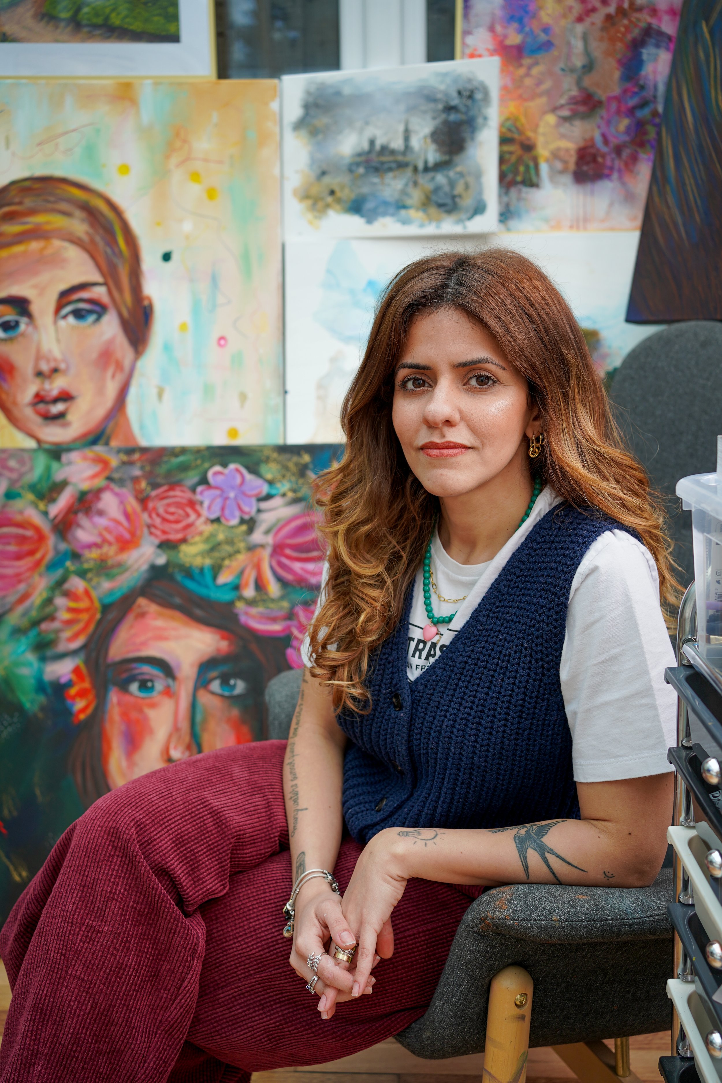 A young woman with long, wavy brown hair and light makeup sitting in an art gallery surrounded by colorful paintings. She is wearing a white T-shirt, a blue knitted vest, and red corduroy pants, with various rings, bracelets, earrings, and a turquoise beaded necklace. She has tattoos on her left arm and is sitting on a chair with her hands resting on it.