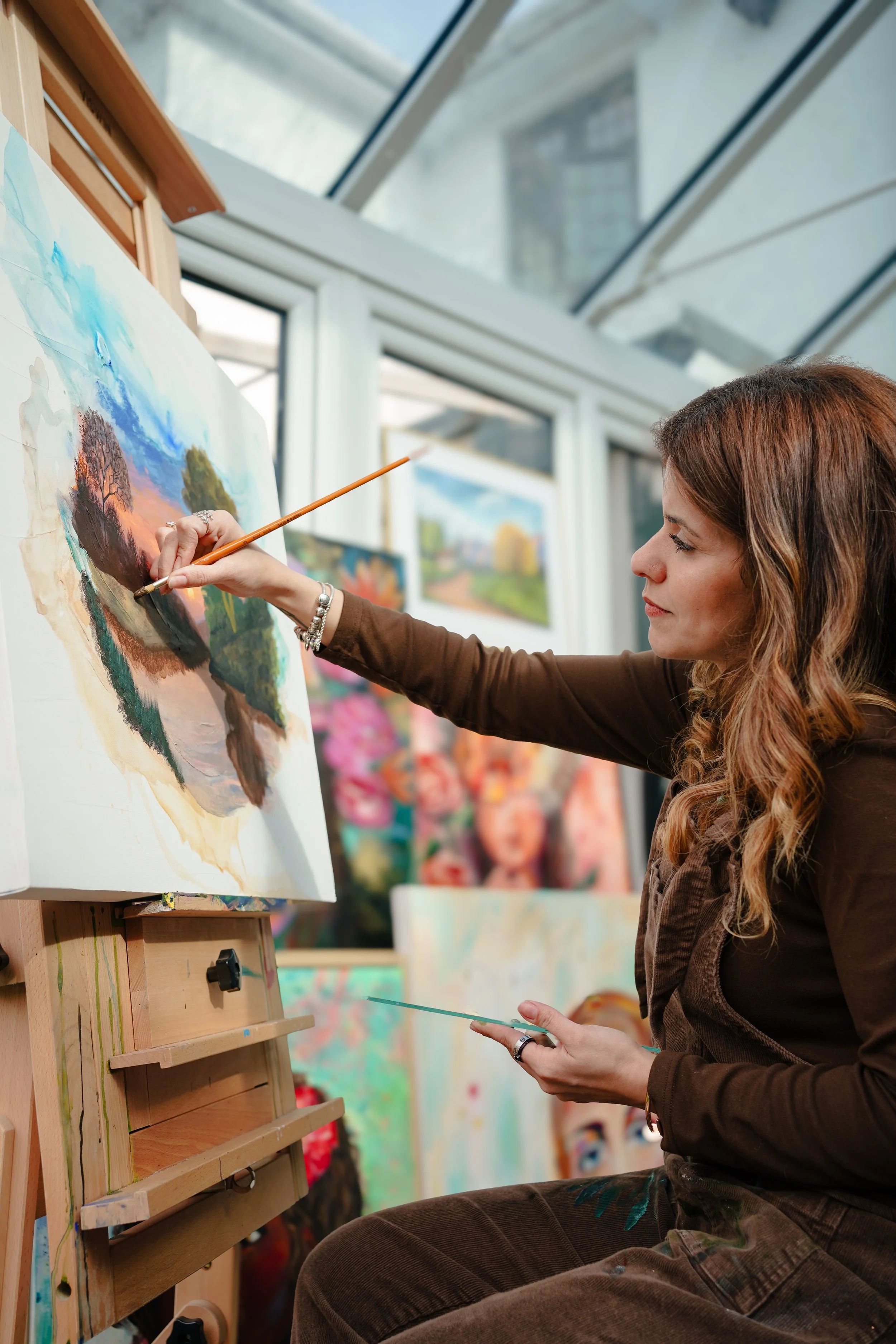 A woman with long, wavy hair is painting a landscape on a canvas in an art studio, with her right hand holding a paintbrush and her left hand holding a palette.
