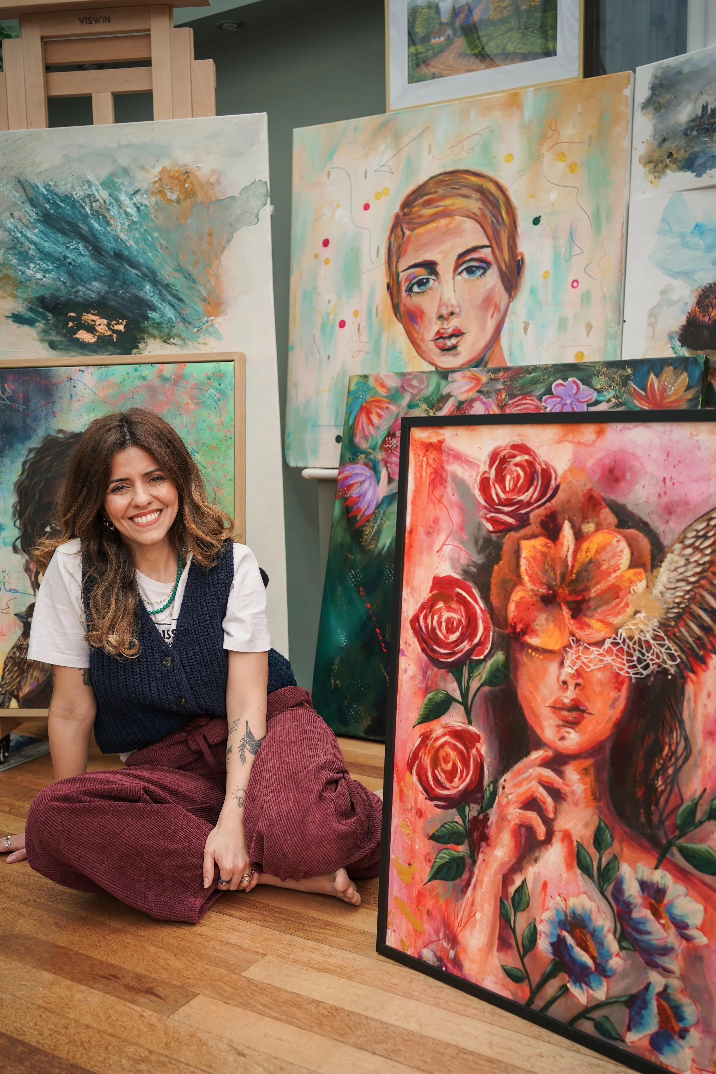 A woman with long wavy brown hair is sitting cross-legged on a wooden floor, smiling. She is wearing a white T-shirt, a dark blue sleeveless vest, and maroon pants. Her right arm has tattoos. Behind her, there are several colorful paintings, includin