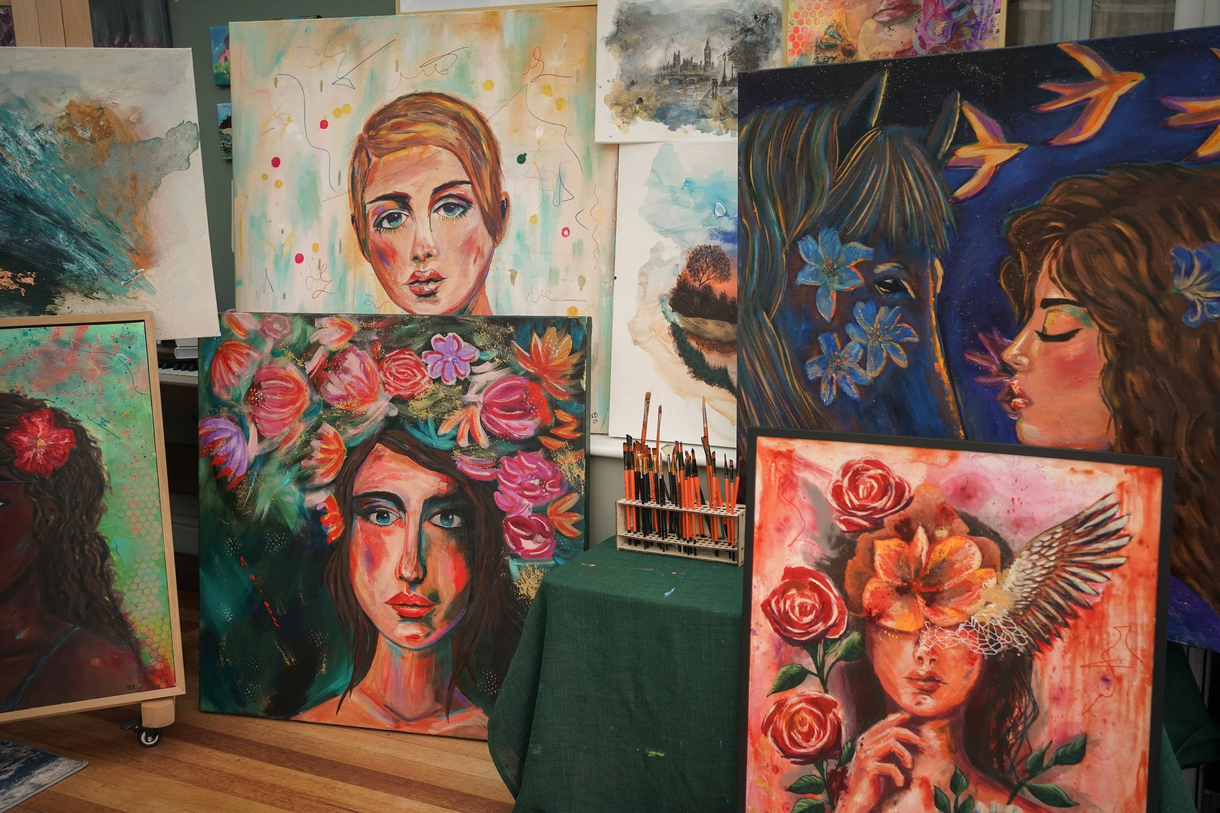 Display of various colorful paintings, including portraits of women, floral arrangements, landscapes, and abstract art, arranged on an easel and the floor.