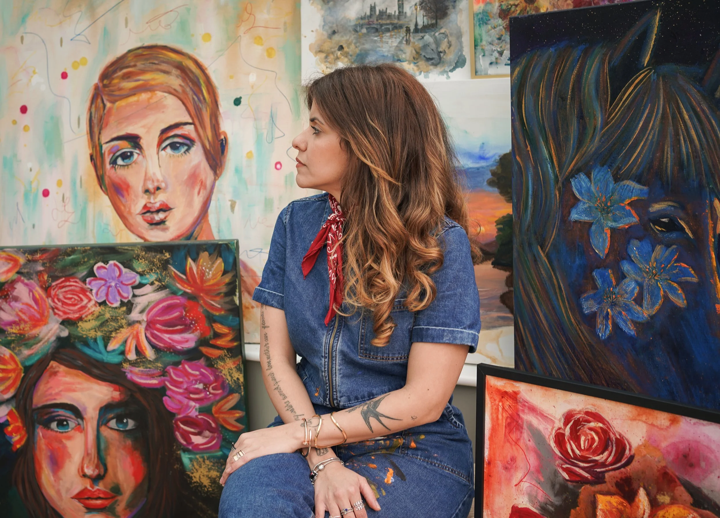 A woman with long, reddish-brown hair and tattoos on her arms, wearing a denim shirt and a red bandana around her neck, sitting in front of and looking at various colorful paintings in an art gallery.