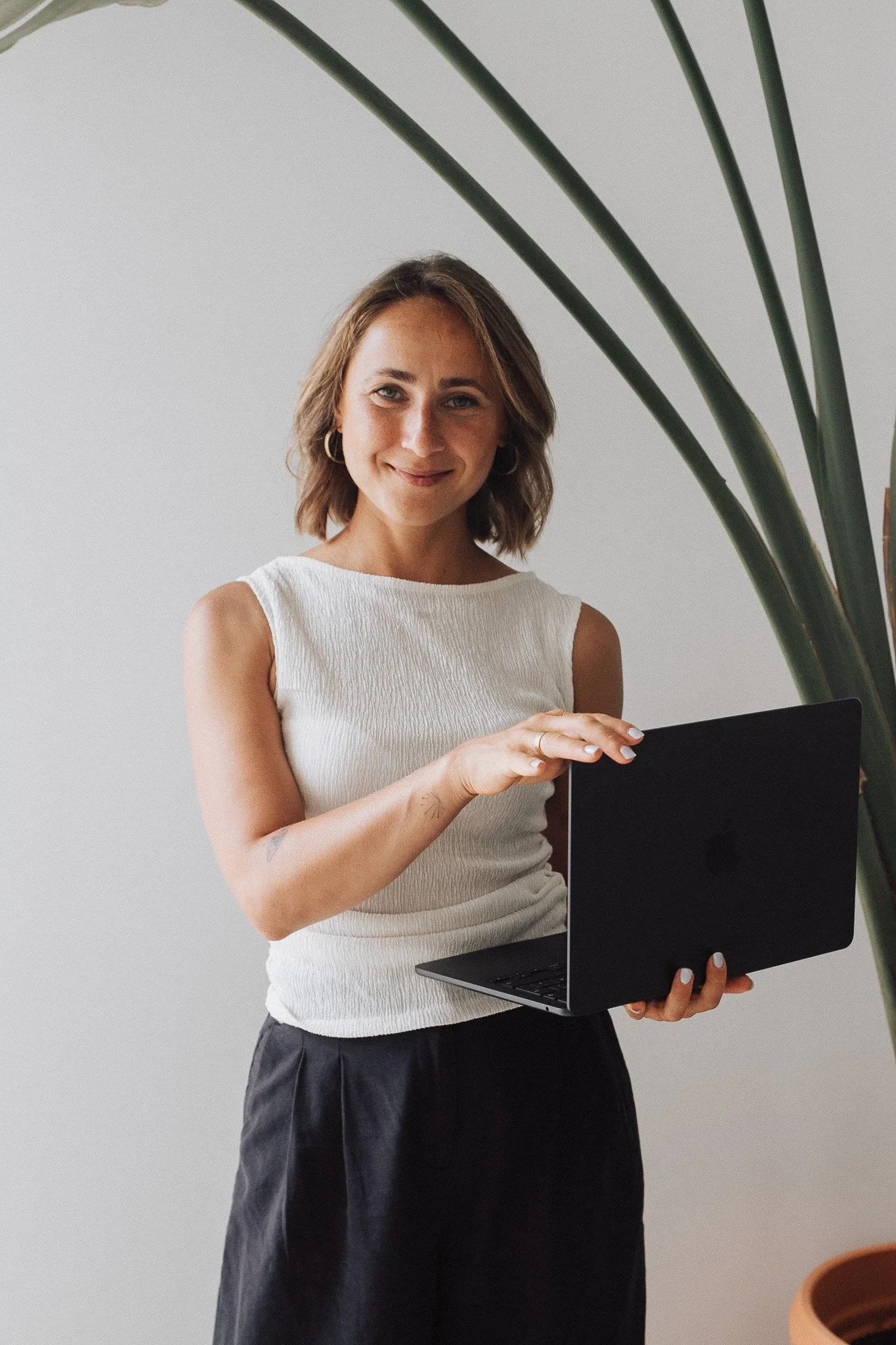 A woman with short brown hair, wearing a white sleeveless top and black pants, is smiling while holding a black laptop. There is a large green plant with long leaves behind her, and the background is a plain light-colored wall.