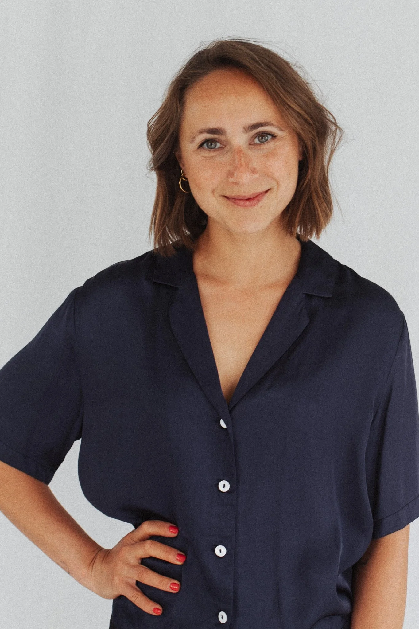 A woman with shoulder-length brown hair, wearing a navy blue button-up shirt and gold hoop earrings, standing with one hand on her hip against a plain light background.
