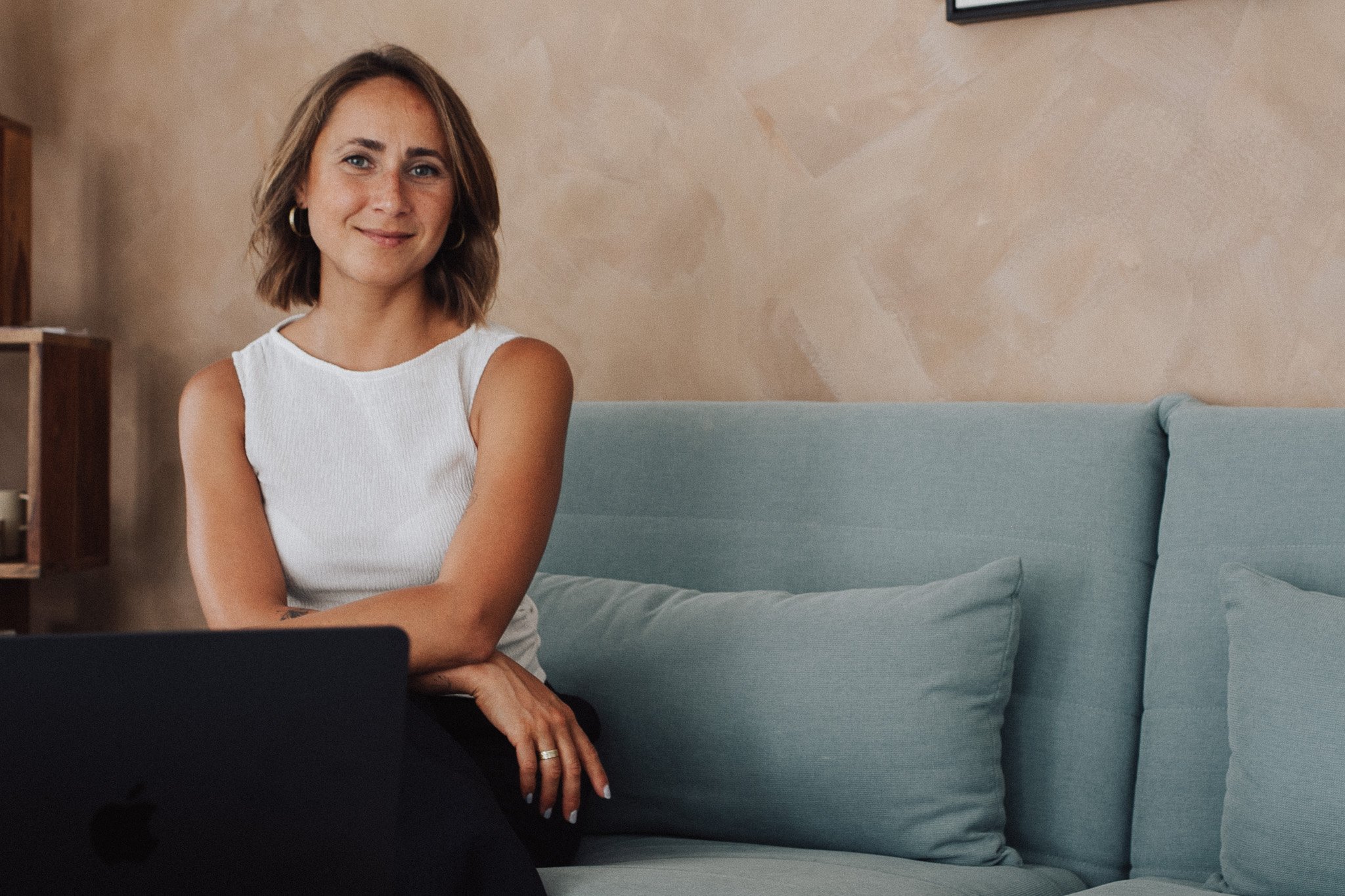Liana sitting on a torquise sofa, brown wall in the background. She is in seated in front of her Laptop, smiling to the camera