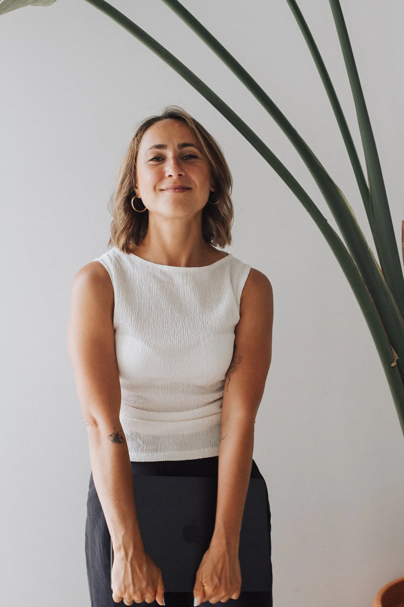 A woman with short brown hair wearing hoop earrings and a white sleeveless top, smiling with eyes closed, standing in front of a plain white wall with large green plant leaves partially visible on the right.
