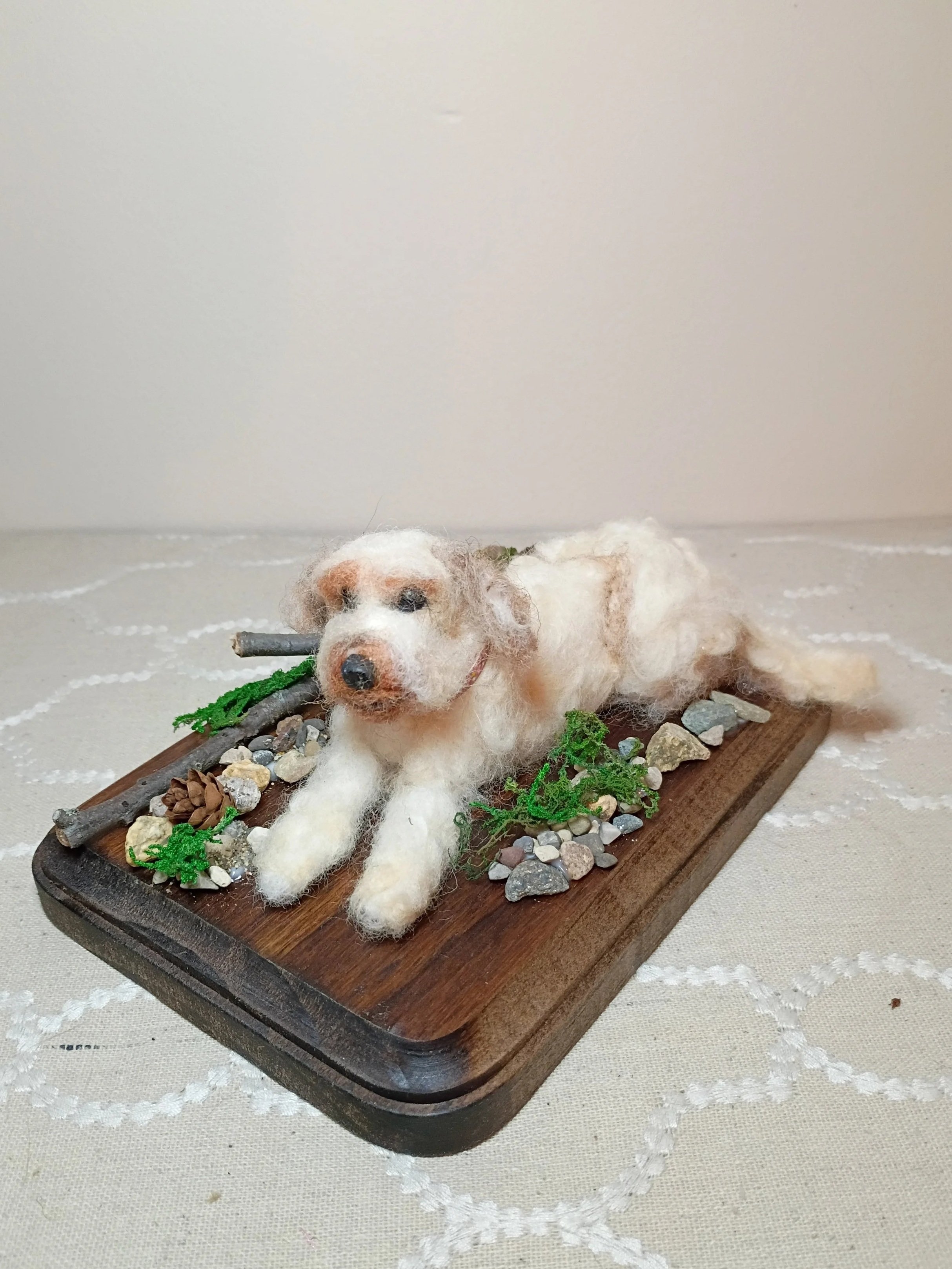 A felted dog with multicolored wool, lying on a wooden tray surrounded by small rocks, twigs, and greenery, on a light-colored table with a subtle pattern.