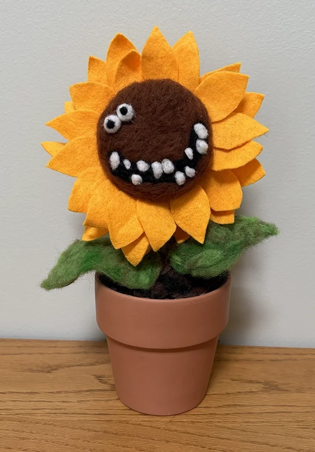 A felt sunflower with a smiley face and googly eyes, potted in a pink flowerpot.