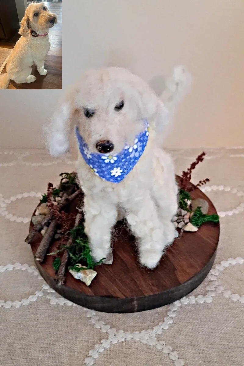 A white stuffed or sculpted dog with a blue bandana decorated with white flowers, sitting on a round wooden platform with small branches, rocks, and green foliage, against a plain wall.
