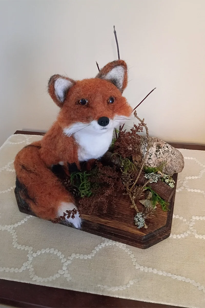 A realistic felted fox sculpture sitting on a wooden display with natural elements like moss, twigs, and a large rock, placed on a table with a beige tablecloth.