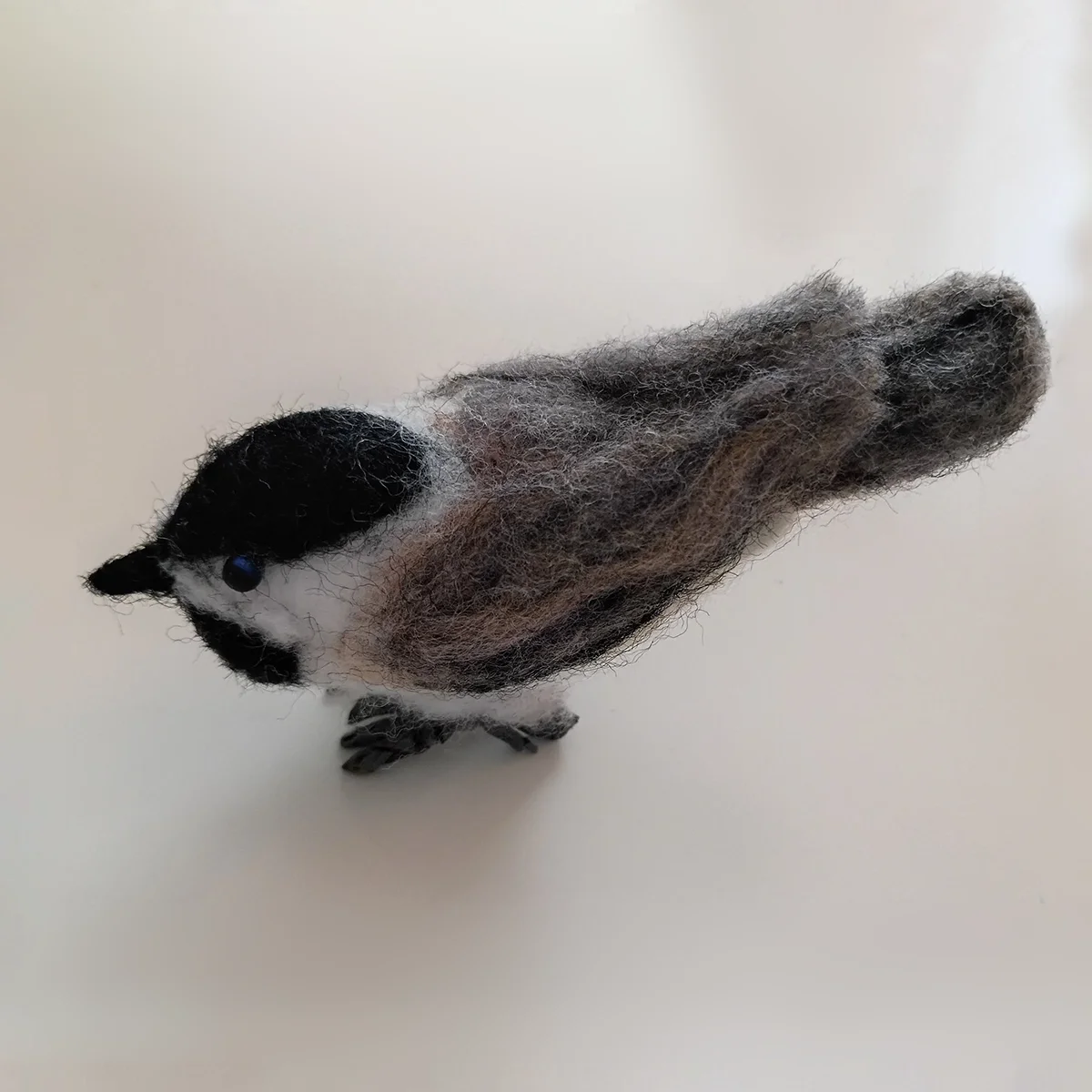 Felted wool craft resembling a small bird with black and white face and gray body, standing on a white surface.